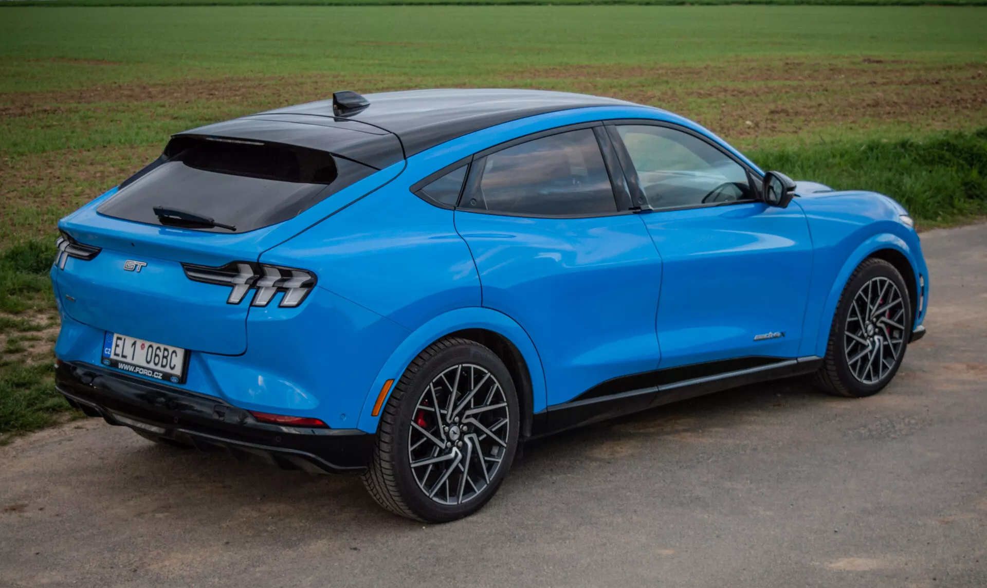 Světle modré elektrické SUV Ford Mustang Mach-E 2020 v dynamickém zobrazení ze tří čtvrtin odzadu zdůrazňující aerodynamický design, elegantní zadní svítilny a sportovní slitinová kola, předvádějící pokročilou technologii a elektrický pohon.