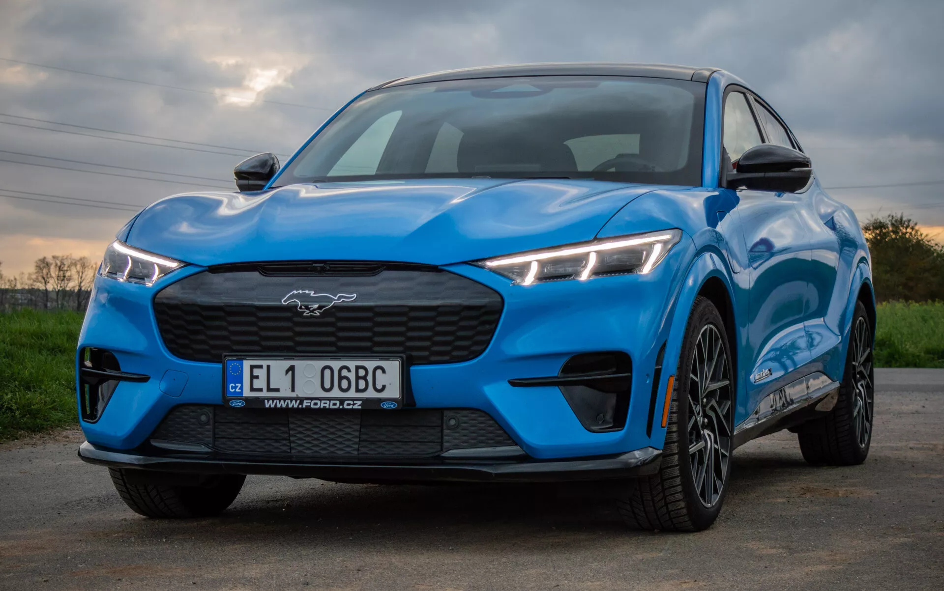 Ford Mustang Mach-E rok 2020 v jasně modré barvě předvádějící sportovní a aerodynamický design z nízkého úhlu pohledu zepředu a ze strany, zdůrazňující úhlová LED světlomety, uzavřenou mřížku chladiče, profilovanou kapotu a kola s vícepaprskovým designem proti přírodnímu pozadí.