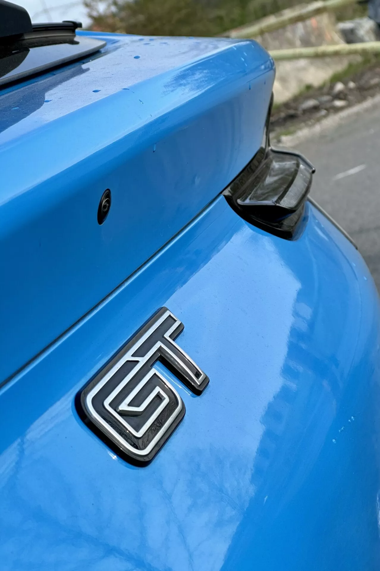 Detailní pohled na zadní část modrého Ford Mustang Mach-E z roku 2020 se zaměřením na kovovou značku GT, zakřivený kufr a aerodynamický zadní spoiler s černým koncovým krytem na mírně rozostřeném pozadí.