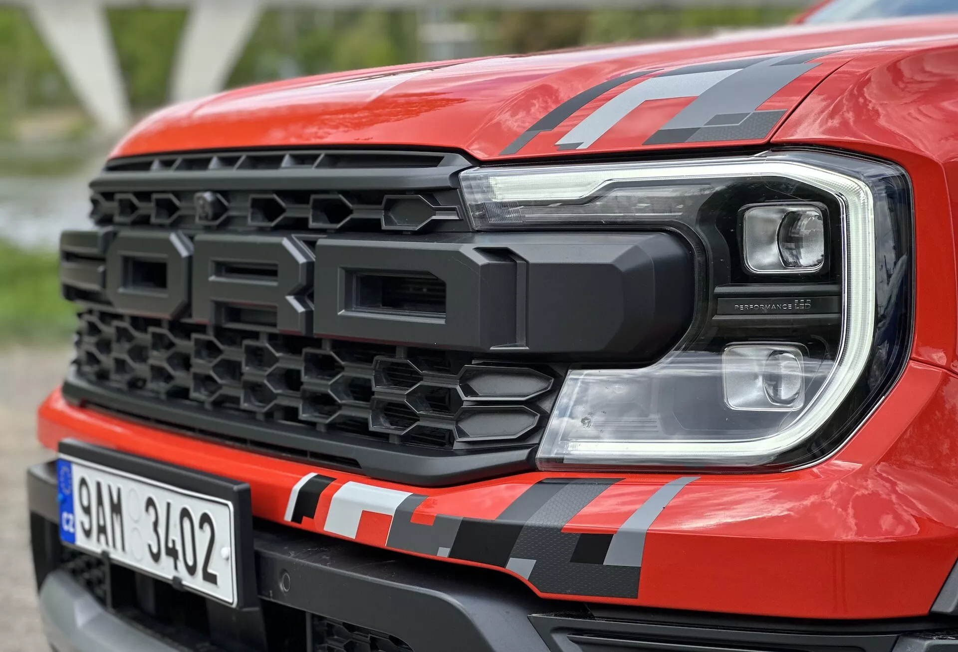 Detailní pohled zepředu na Ford Ranger 2022, zvýrazňující černou mřížku s hexagonálními vzory a moderní design LED světlometů s červenými pruhy na kapotě a doplňkovým světelným prvkem, předvádějící pokročilou technologii a sportovní estetiku