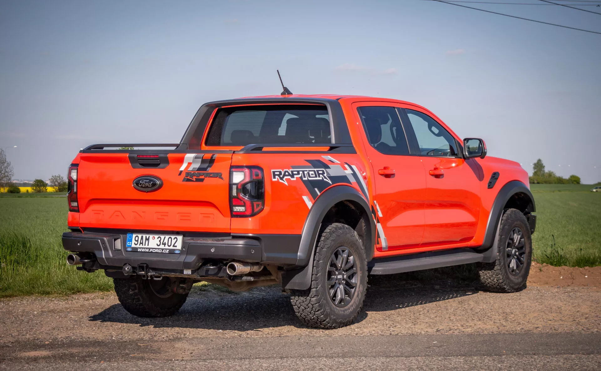 Světle oranžový pickup Ford Raptor roku 2022 z bočního zadního úhlu, venku na poli, předvádějící jeho schopnosti pro jízdu v terénu s vysokou světlou výškou, dveřmi double cab, velkými černými rozšířeními podběhů, černými koly s více paprsky a terénními pneumatikami, a zadní značení 'RAPTOR' na víku nákladového prostoru.