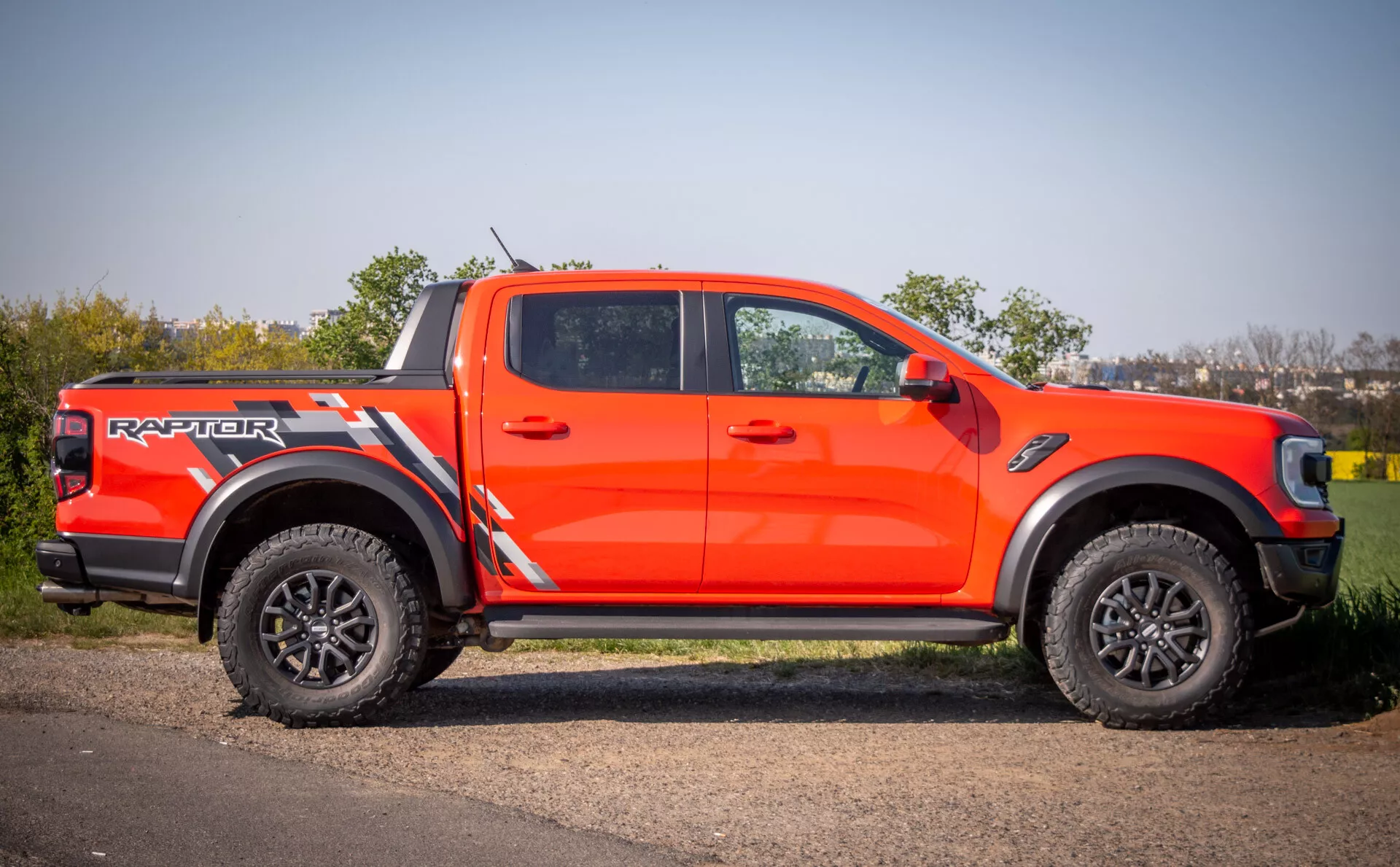 2022 Ford Ranger Raptor v jasně oranžové barvě předvádějící agresivní styling, terénní schopnosti s velkými černými ráfky a pneumatikami s hlubokým profilem, parkovaný na dlážděné ploše vedle trávy během dne