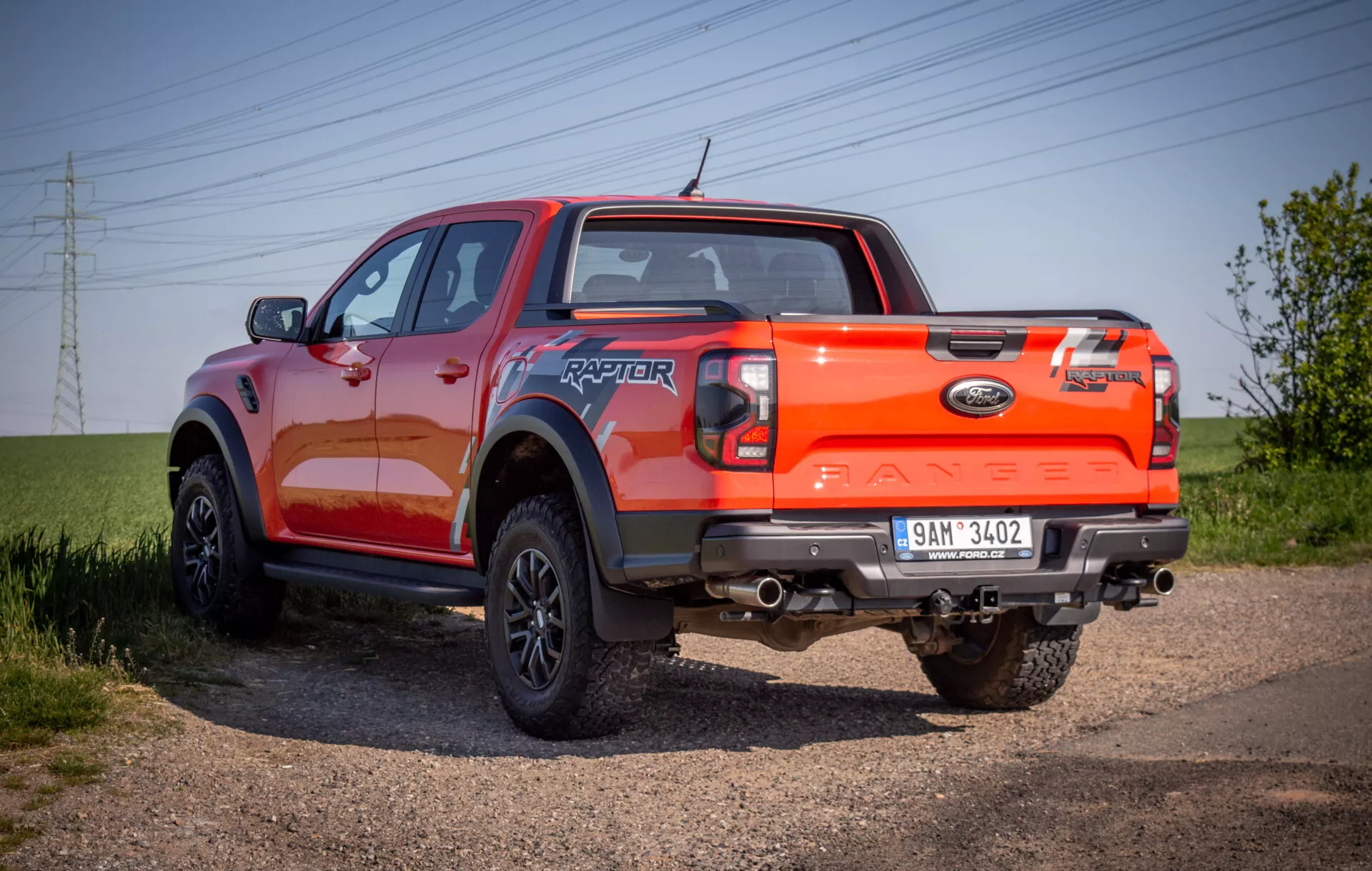Pickup Ford Ranger Raptor 2022 vyobrazený na štěrkové cestě, prezentující jeho robustní zadní tříčtvrteční úhel s důrazem na schopnost terénní jízdy a výraznou oranžovou barvu laku.