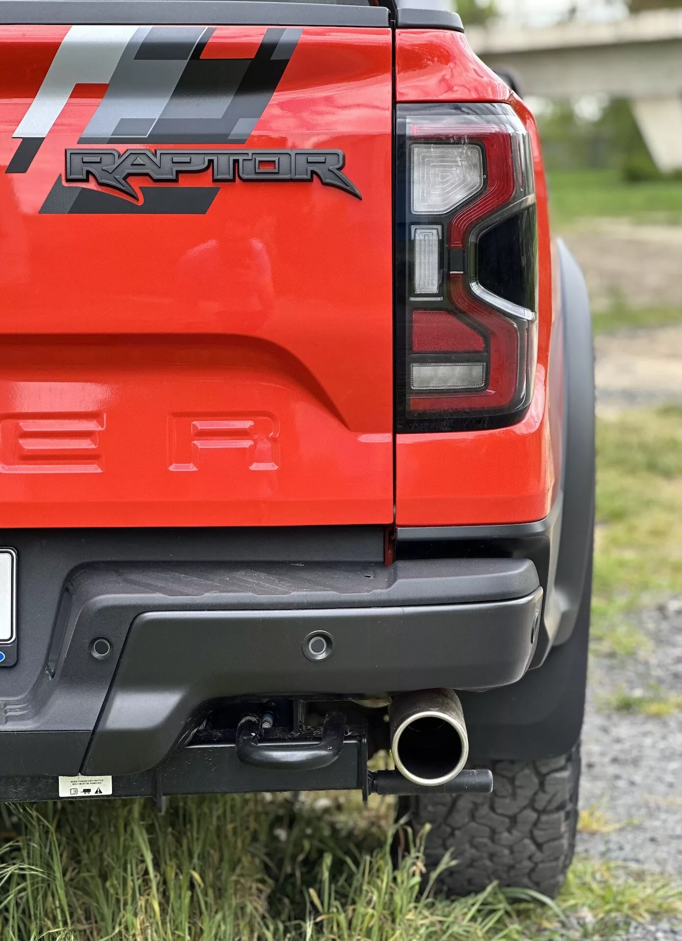 Detailní pohled na zadní roh červeného Fordu Ranger Raptor 2022 s LED zadním světlem, černým zadním nárazníkem se šedým panelem, tažným zařízením, leštěnou koncovkou výfuku a terénní pneumatikou, zdůrazňující jeho schopnosti v terénu a designové detaily.