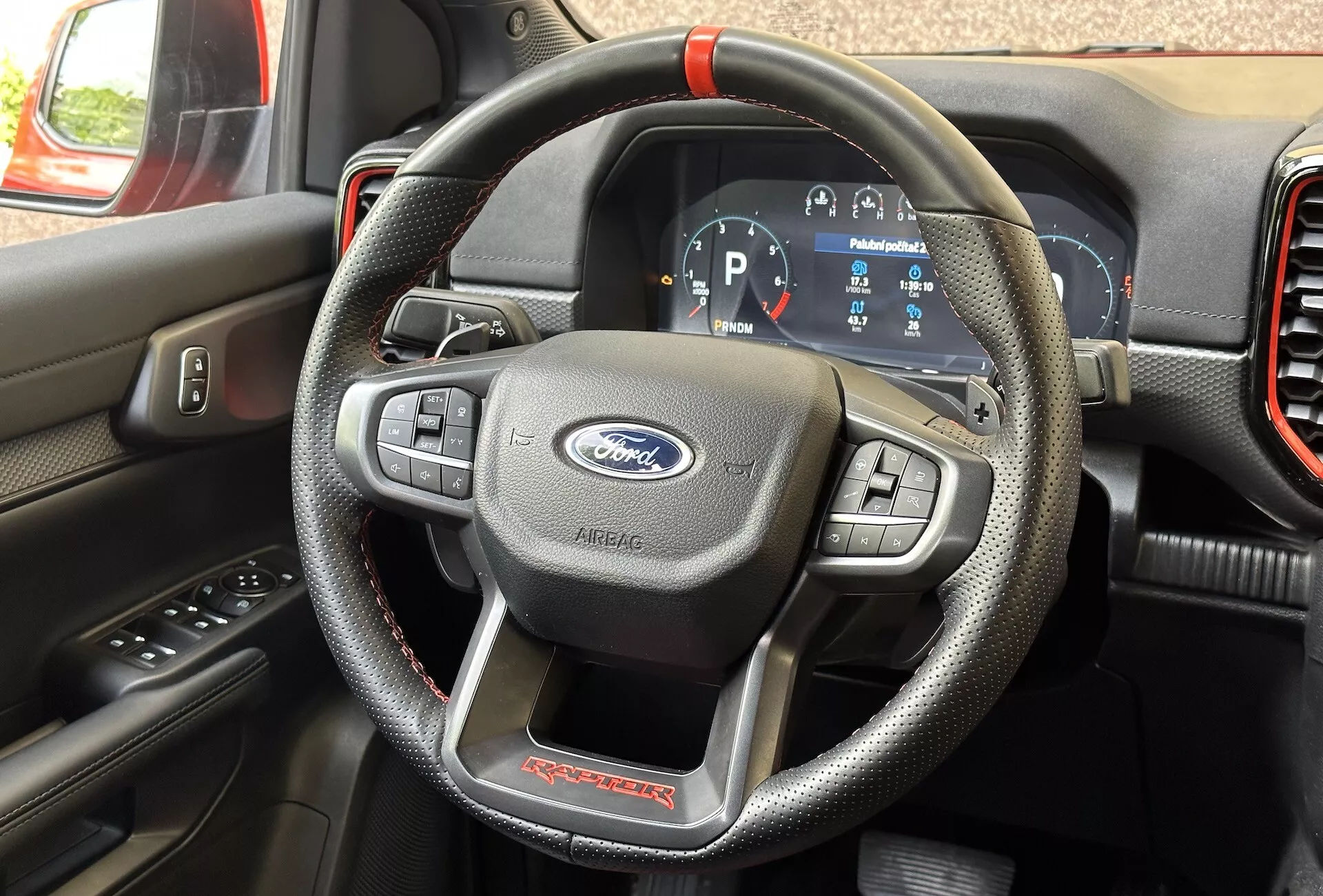 Podrobný interiérový pohled na Ford Ranger Raptor 2022 zaměřený na volant připomínající kůži s červeným prošíváním a multifunkčními ovládacími prvky a digitální přístrojovou desku ukazující vozidlo v režimu Park.