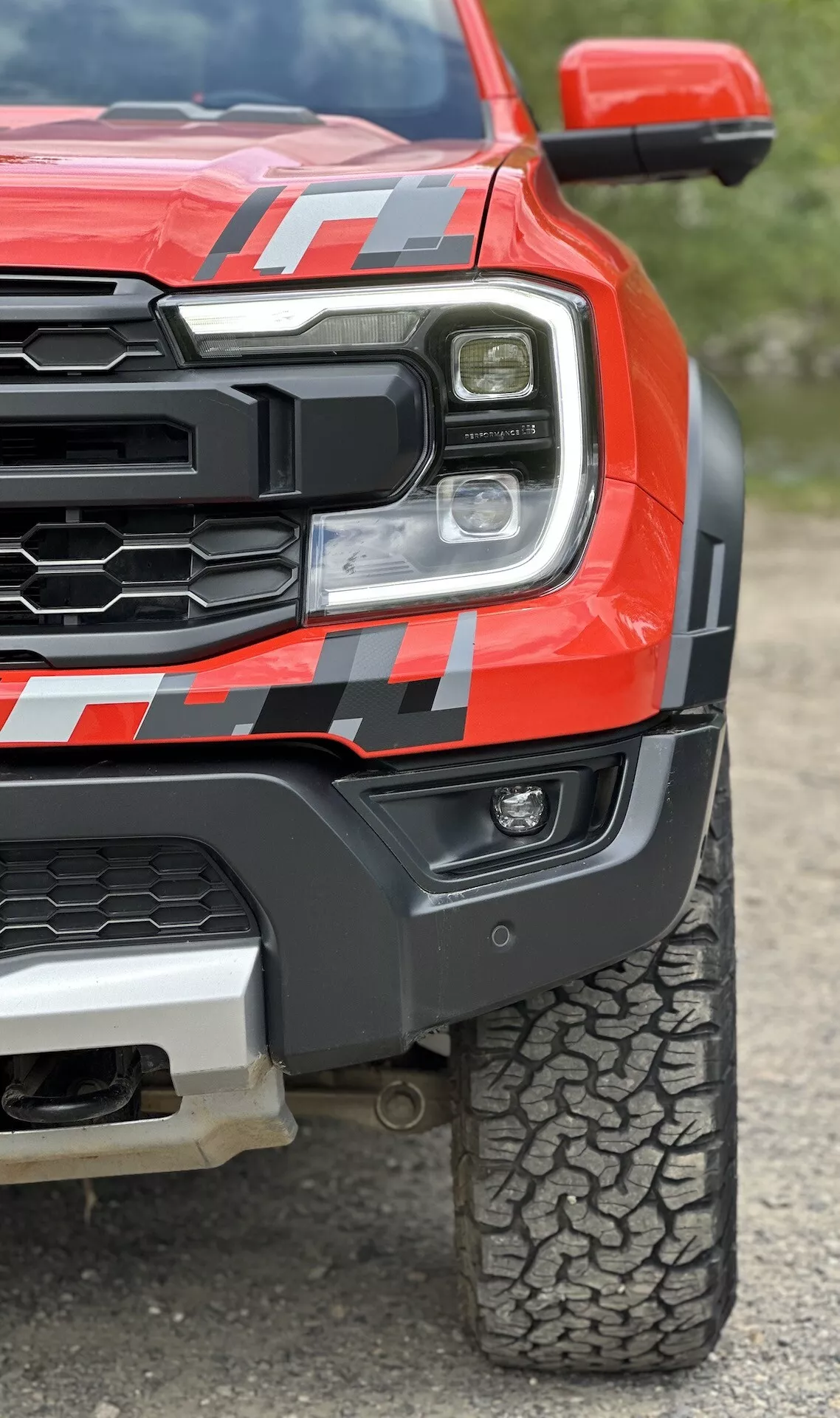 Detailní pohled na červený pickup Ford Ranger 2022, zobrazující LED světlomet, robustní mřížku, svalnaté přední blatníky s dekálem a vzor dezénu pneumatiky pro terénní použití z nízkého úhlu pro zdůraznění odolnosti a pokročilé technologie