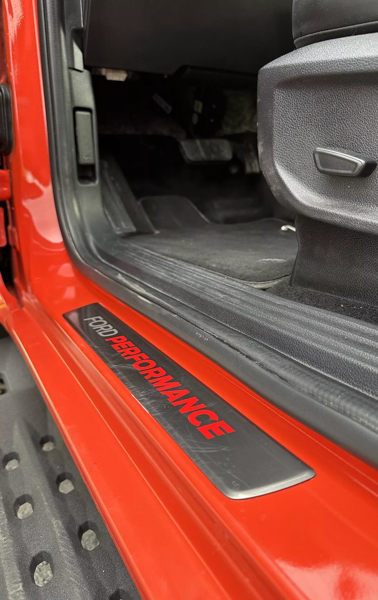 Detailní pohled na prah dveří Ford Ranger 2022 s odznakem 'FORD PERFORMANCE', lesklý červený exteriér, černý vnitřní koberec, sedadlo z šedé látky se sportovním designem a odolnou černou gumovou podlahovou rohoží