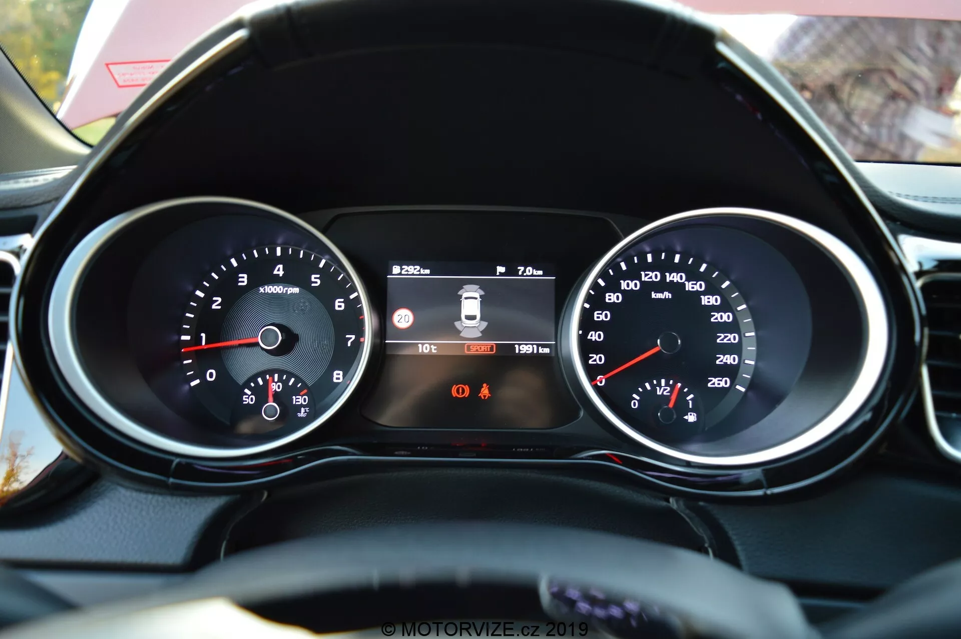 Detailní pohled z pohledu řidiče na přístrojový panel Kia Ceed 2021 a část volantu s koženým povrchem. Funkce zahrnují tachometr vlevo s hodnotami RPM, centrální digitální displej zobrazující systém varování při opuštění jízdního pruhu, čas a počítadlo kilometrů a rychloměr vpravo s rychlostí v km/h. Pod centrálním displejem jsou viditelné dvě výstražné kontrolky, přičemž kontrolka připomínající připoutání bezpečnostních pásů je identifikovatelná.