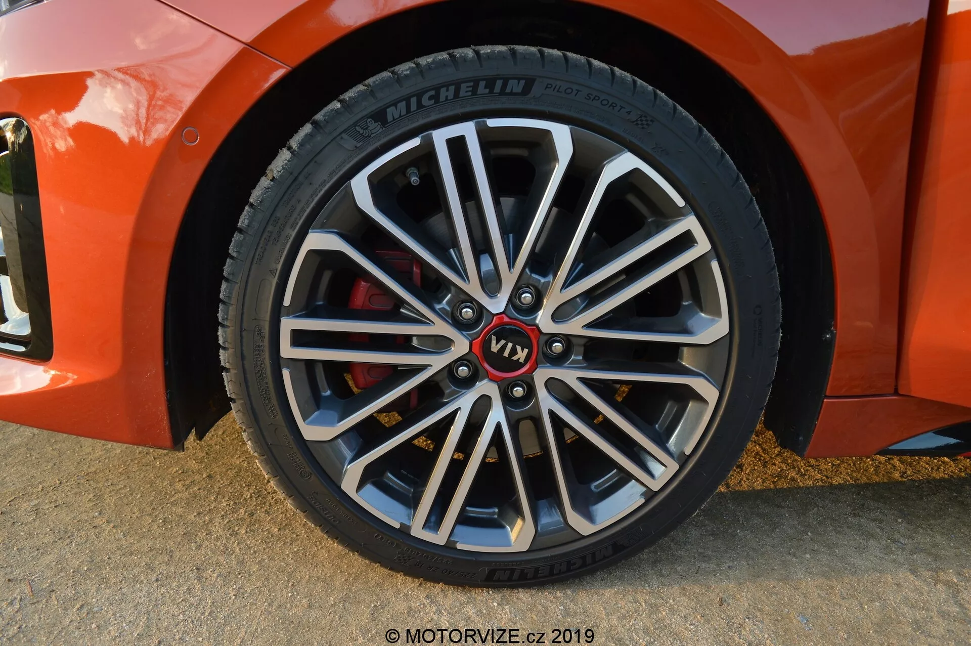 Detailní záběr předního kola z roku 2021 Kia Ceed na straně spolujezdce, mírně diagonální úhel, ukazuje vícepaprskové alu kolo s tmavšími vnitřními sekcemi a jasnými opracovanými okraji. Disponuje značkovaným středem s červeným označením 'VT1', pneumatikou Michelin s podrobnými informacemi na boční stěně a červeným brzdovým třmenem, což naznačuje komponenty orientované na výkon. Měkké stíny a zvýraznění naznačují, že fotografie byla pořízena v přirozeném osvětlení, pravděpodobně během zlaté hodiny.