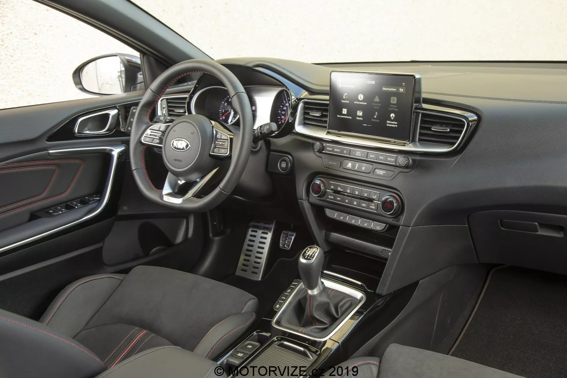 Interiér Kia Ceed 2021, zaměřený na stranu řidiče z boční perspektivy. Volant s rozpoznatelným logem výrobce obsahuje multifunkční ovladače pro správu infotainmentu, tempomatu, telefonního připojení a možná i hlasových příkazů. Za volantem jsou vidět rychloměr, otáčkoměr a měřidla řidičských informací. Středová konzole má dotykovou obrazovku infotainmentu, která nabízí navigaci, nastavení vozidla, přehrávání médií a připojení Bluetooth. Níže jsou manuální ovladače pro audio a klimatizační systémy. Řadicí páka manuální převodovky, obklopená tlačítky pro jízdní režimy nebo kontrolu trakce, a sportovní sedadla s koženými a textilními vložkami, zvyšují prémiový, výkonnostně orientovaný pocit. Kovové pedály v prostoru pro nohy řidiče naznačují důraz na výkon, zatímco detaily s chromovou úpravou na ventilech klimatizace, klikách dveří a dalších detailech přidávají sofistikovanost. Dveřní panel obsahuje ovladače elektrických oken a možná i ovladače elektrických zpětných zrcátek, a je viditelná část zpětného zrcátka, která naznačuje funkce jako automatické stmívání nebo integrovaný kompas.