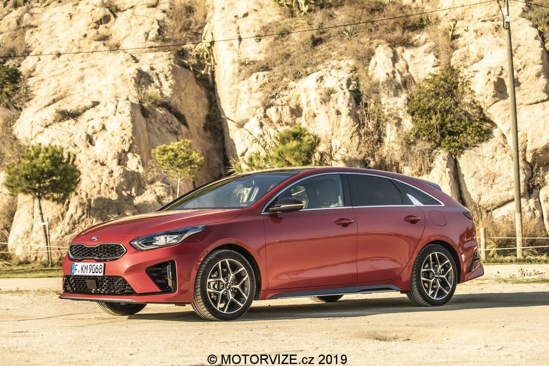 Boční profil červeného hatchbacku Kia Ceed 2021, s elegantním, aerodynamickým designem se sklánějícími se liniemi střechy a konturovanými liniemi na dveřích. Pět dveří, chromové okenní lišty a začerněné B-sloupky vytvářejí efekt nepřetržitého skleněného panelu. Vícepaprsková litá kola s viditelnými kotoučovými brzdami dodávají sportovní nádech. Ostré světlomety a částečně viditelná mřížka označují předek, zatímco zrcátka v barvě karoserie s integrovanými směrovými světly zlepšují viditelnost a bezpečnost.