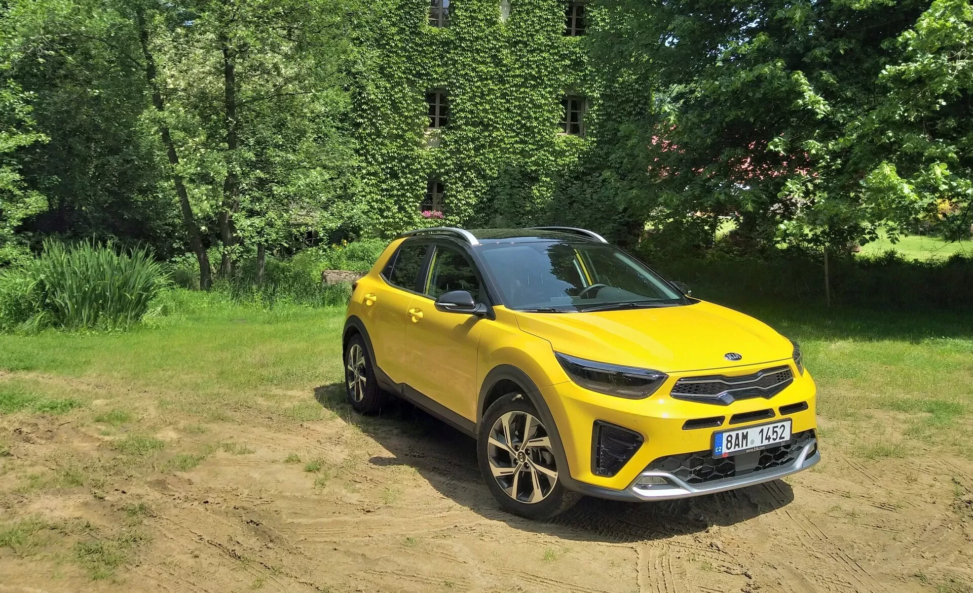Vibrantní žluté crossover SUV Kia Stonic 2017 ve tříčtvrtečním pohledu zepředu zdůrazňující jeho přední mřížku, LED světlomety, kapotu a stranu řidiče, umístěné proti přírodnímu pozadí se slunečním světlem na čisté půdě, předvádějící schopnosti pro jízdu v lehkém terénu, sportovní slitinová kola a moderní designové prvky.