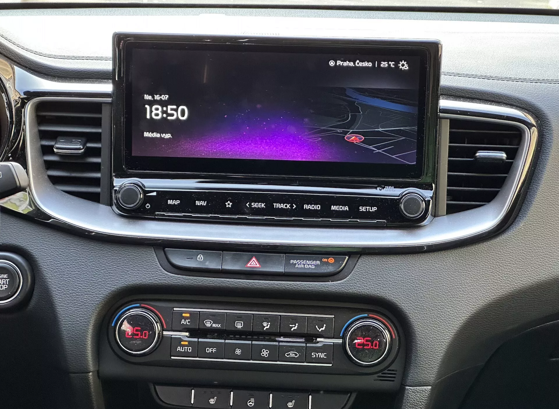 Detailní pohled na interiér Kia Xceed 2022 ukazující středovou konzoli s dotykovou obrazovkou infotainmentu, která zobrazuje barevné rozhraní s navigací a informacemi o médiích, řadu tlačítek pro rychlý přístup, ovládání výstražných světel a panel s dvojzónovou klimatizací, vše orámováno stříbrnou lištou.