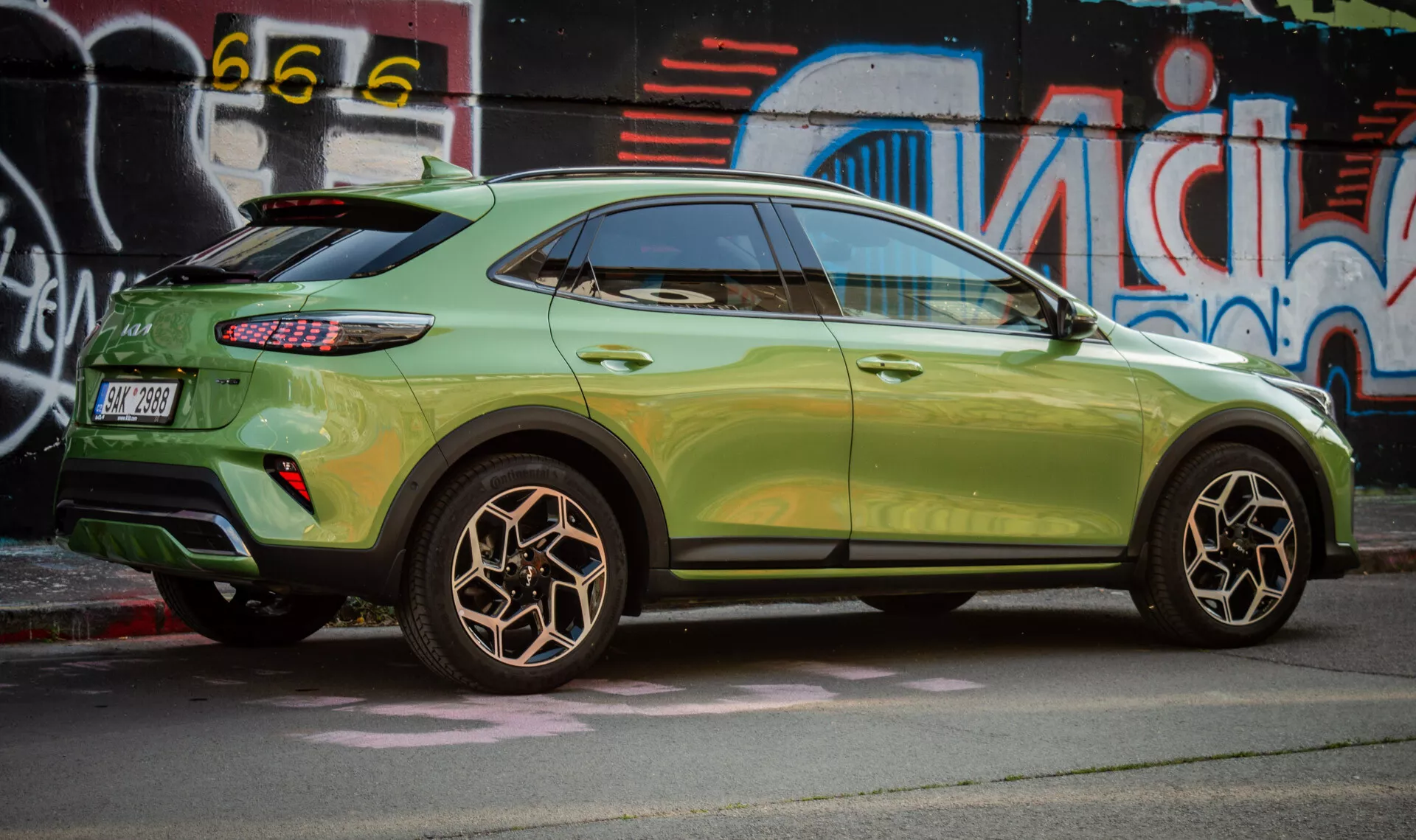 Pohled z boku na Kia XCeed 2022 zdůrazňující design zadního kola, metalickou zelenou barvu, LED zadní světla a stylizaci kompaktního SUV s aerodynamickými prvky na pozadí městského street artu.