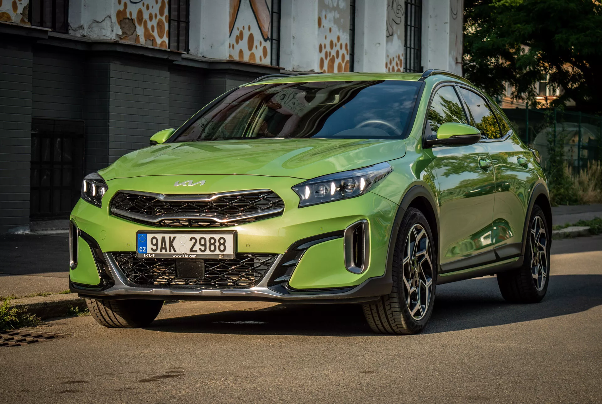Kia XCeed 2022 ve výrazné limetkově zelené barvě s LED světlomety, výraznou přední maskou s emblémem značky, sportovním předním nárazníkem a velkými slitinovými koly, umístěná na zpevněné cestě s městským pozadím