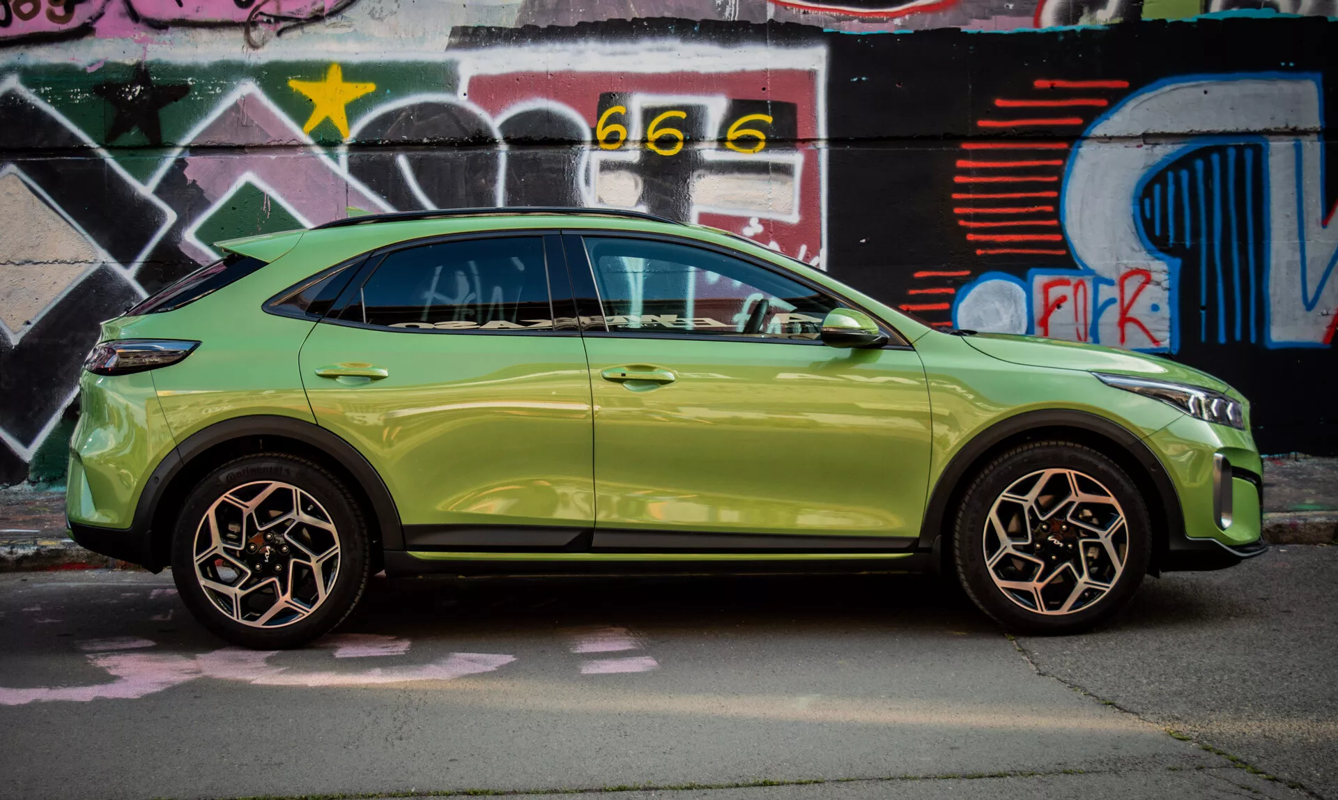 Kompaktní SUV Kia XCeed 2022 v limetkově zelené barvě zaparkované u zdi pokryté graffiti, předvádějící svůj elegantní design a pokročilé funkce, jako je bezklíčové vstupní systém a integrované směrové signály v bočních zrcátkách.
