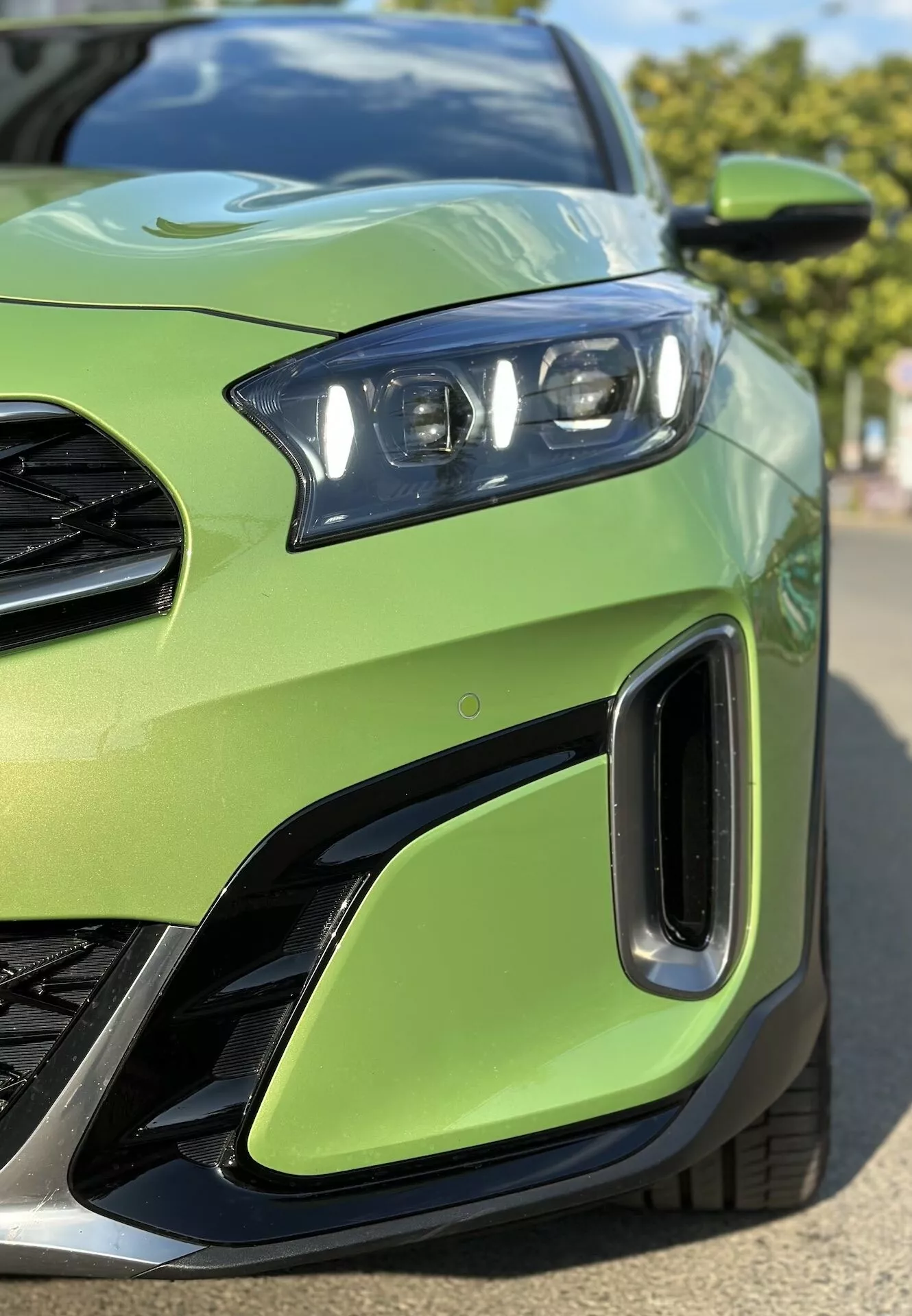 Detailní záběr na přední levou část Kia Xceed 2022 ukazující sestavu LED světlometů, část mřížky, vzduchové vstupy na předním nárazníku, proti limetkově zelené barvě karoserie, zdůrazňující sportovní design vozidla a pokročilou světelnou technologii.