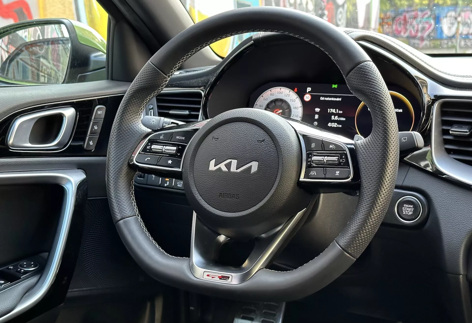 Interiér Kia XCeed 2022 z pohledu řidiče s černým volantem potaženým kůží s multifunkčními ovládáními a odznakem GT line, digitálně-analogovým přístrojovým panelem, částí středové konzoly s ventilačními otvory, ergonomickým designem palubní desky a panelu řidičových dveří s ovládáním, odrážející městské prostředí