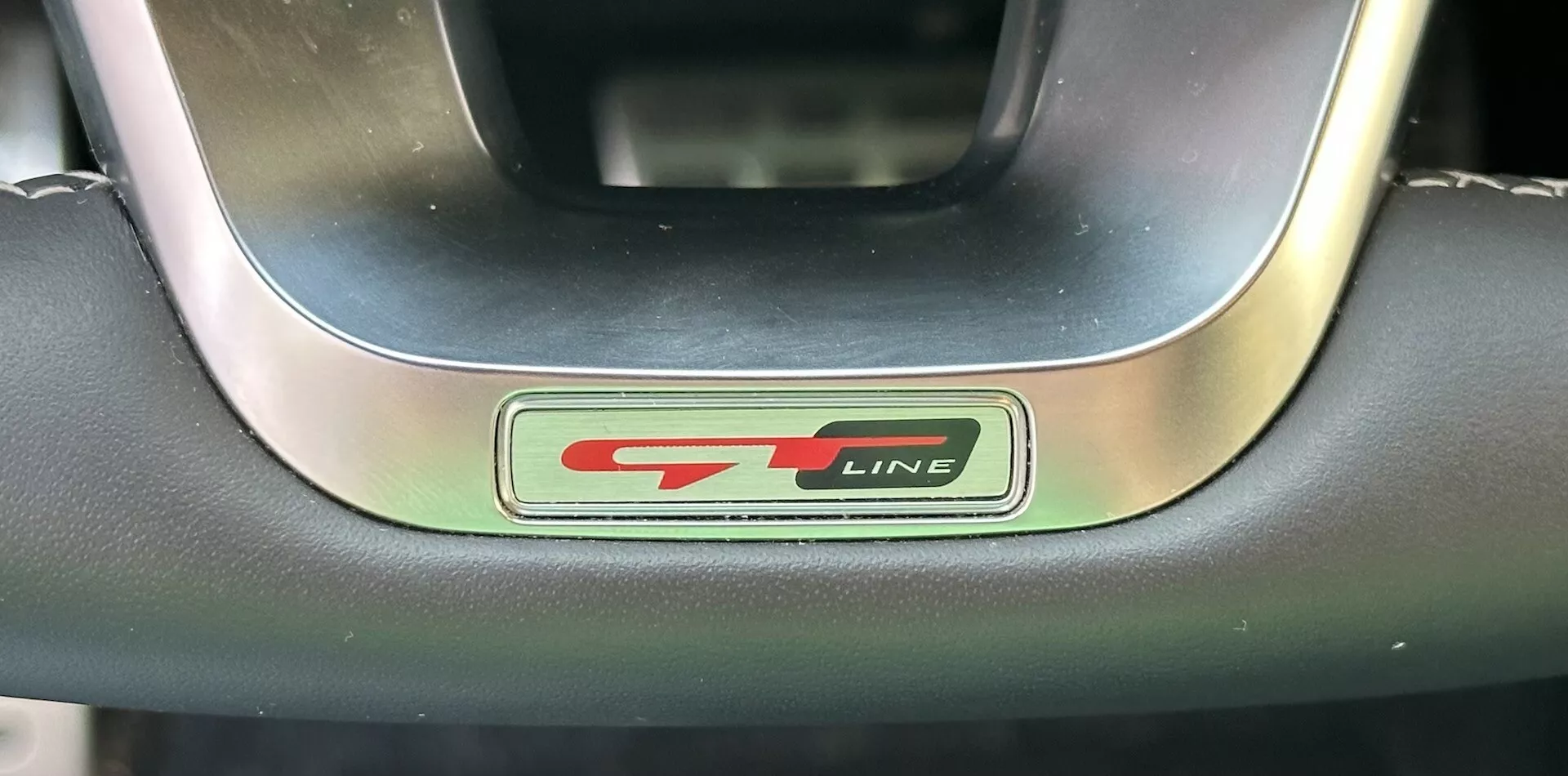 Detailní záběr na emblém volantu Kia XCeed 2022 GT-Line s kovovým a černým písmem a sportovním červeným akcentem, obklopeným prémiovým koženým a kovovým volantem