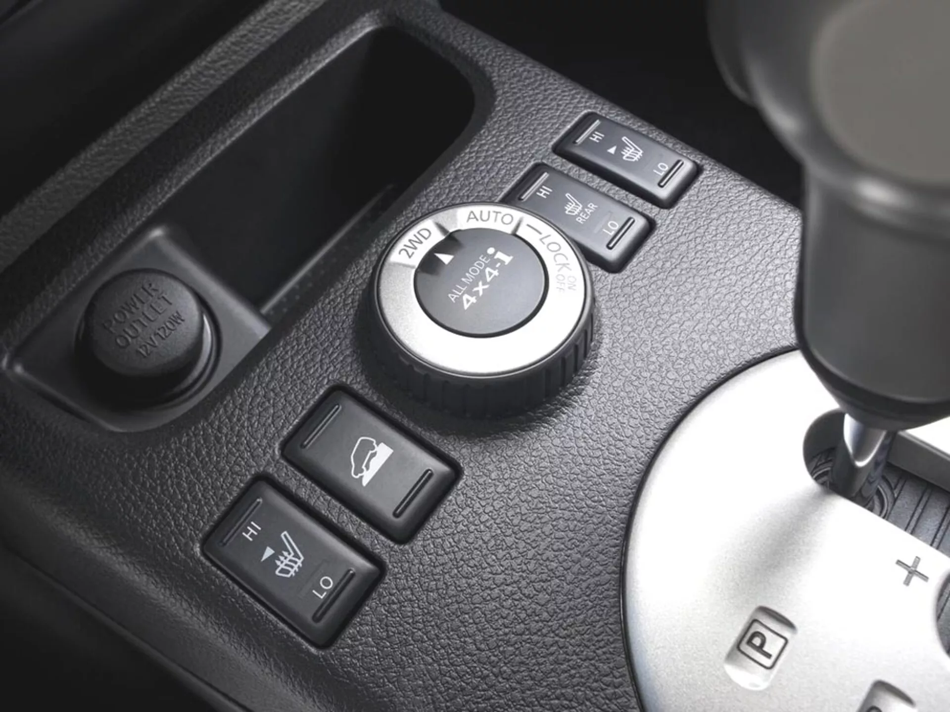Detailní pohled na středovou konzolu Nissanu X-Trail 2010 s ovládacím kolečkem 4WD, tlačítky pro vyhřívání sedadel, tlačítkem pro zásuvku a řadicí pákou s možností manuálního řazení