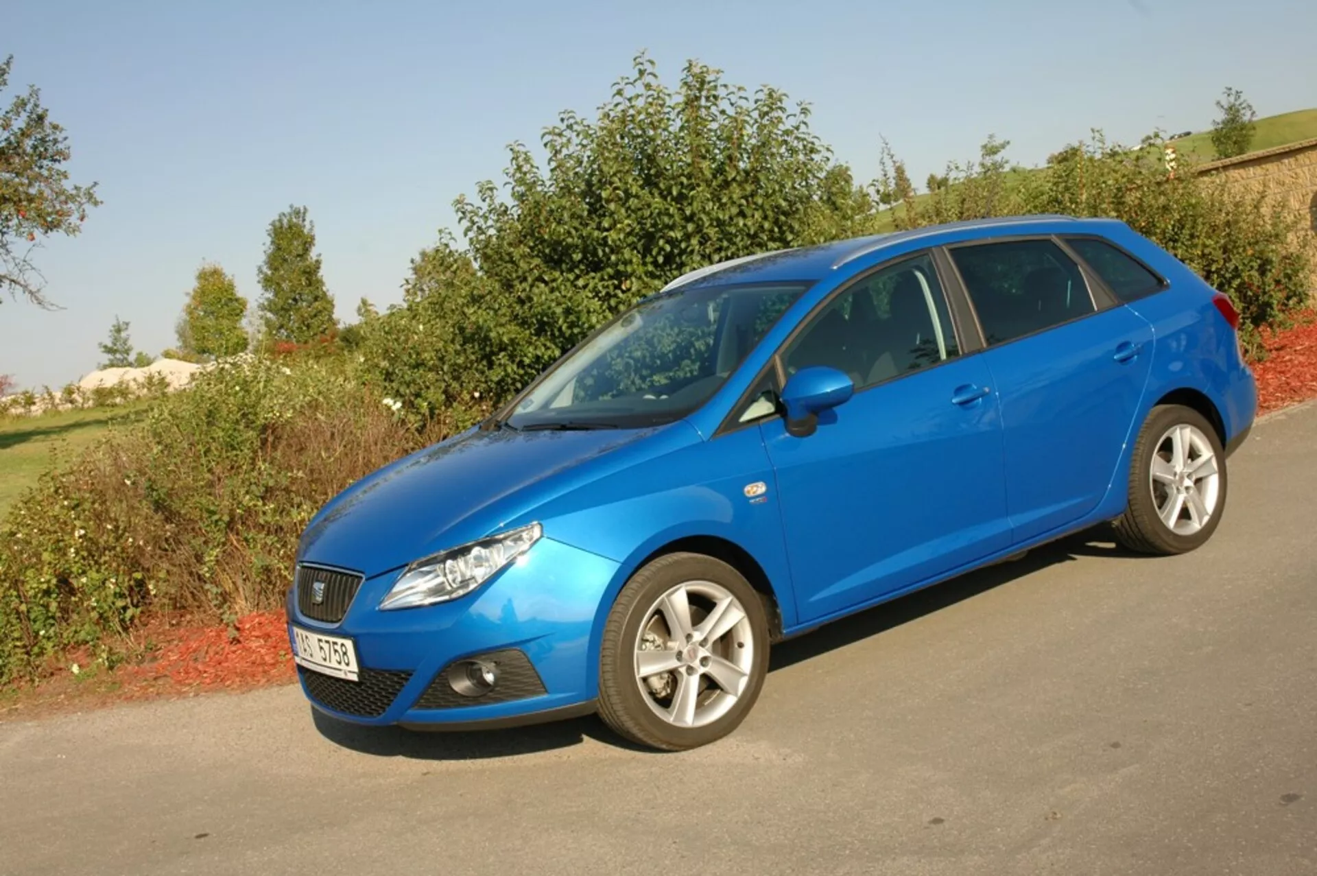 Světle modrý hatchback Seat Ibiza 2012 zaparkovaný na asfaltu proti zeleni, předvádí svou aerodynamickou přední mřížku, zpětně natočené světlomety a boční profil s vícepaprskovými slitinovými koly pod přirozeným denním světlem
