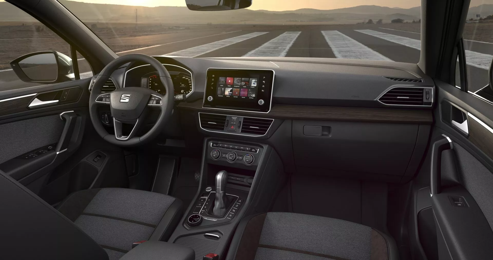 Interiér Seat Tarraco 2019 z pohledu zadního sedadla, zobrazující multifunkční volant, dotykový displej infotainmentu ve středu palubní desky, tlačítka klimatizace, páku automatické převodovky a sedadlo cestujícího potažené tkaninou, pod jemným osvětlením interiéru