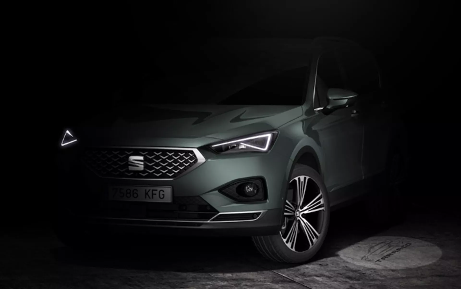 Seat Tarraco 2019 prezentováno v temně osvětlené scéně s důrazem na jeho elegantní přední část, geometrickou mřížku, LED světlomety s výraznými denními světly, a modelovanou kapotu, společně s detailním pohledem na slitinové kolo, zdůrazňující luxusní a moderní technologické prvky vozu