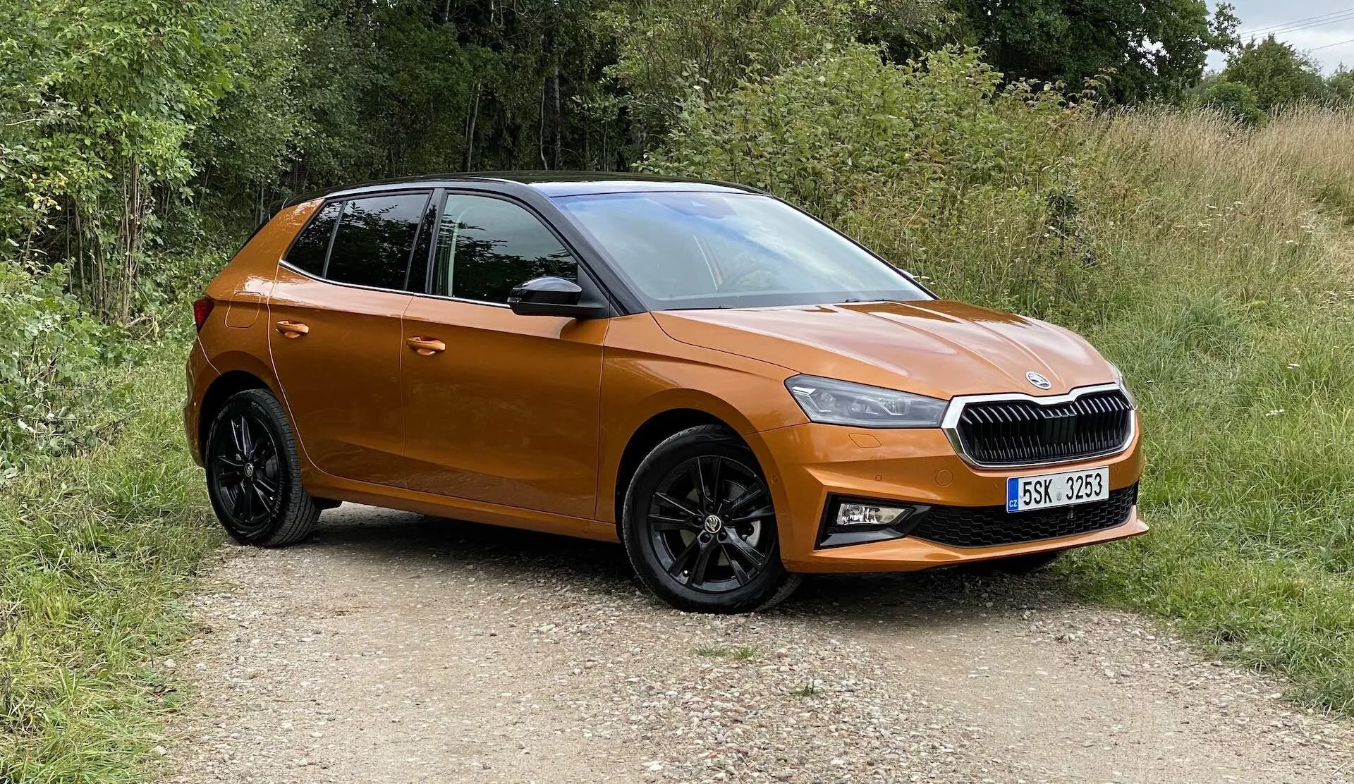 Obrázek moderního hatchbacku Škoda Fabia 2021, zachyceného z tříčtvrtečního předního pohledu, ukazujícího přední levý bok, dveře řidiče, levý blatník a část střechy. Vozidlo je na nezpevněné cestě obklopené zelení, což znamená venkovské prostředí. Viditelné prvky zahrnují vodorovnou mřížku s centrálním emblémem, ostré LED světlomety, metalický oranžový lak, boční zrcátka lakovaná ve stejném odstínu, černá vícepaprsková kola z lehkých slitin s dezénovanými pneumatikami a konturované dveře s elegantními liniemi. Auto vypadá nové, kompaktní a neupravené.