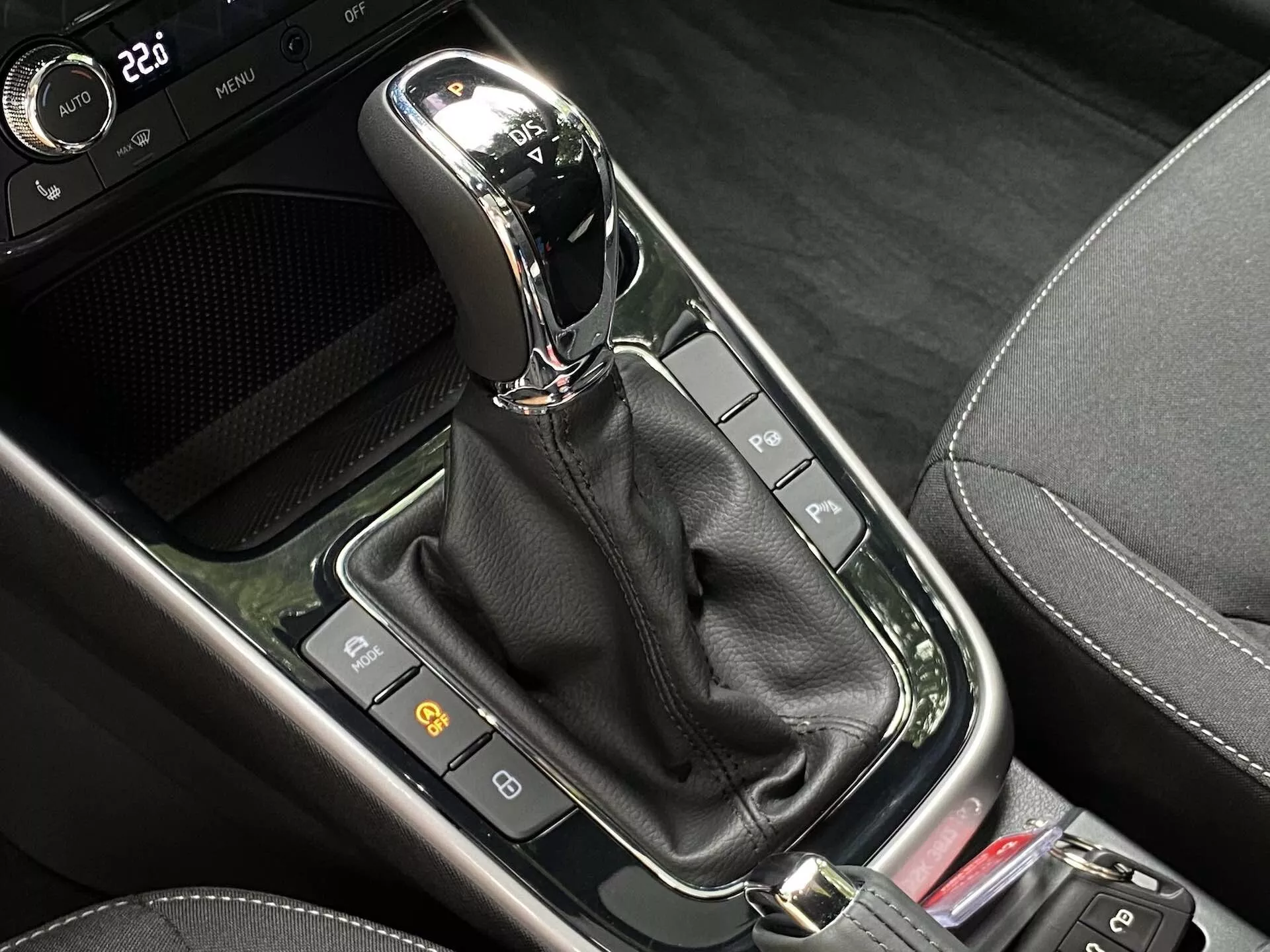 Detailní pohled na automatickou řadicí páku ve středové konzoli vozu Škoda Fabia 2021. Moderní řadicí páka, umístěná svisle v režimu 'P' (parkování), je opatřena chromovým nebo kovovým povrchem na horní části a je zasazena do lesklého černého panelu. Pod ní je černý kožený řemínek ukrývající mechanismus. Kolem řadicí páky je řada tlačítek a indikátorů, včetně dvou vlevo, které se pravděpodobně týkají bezpečnostních funkcí vozu, a části červeného tlačítka vpravo. Oblast kombinuje různé černé materiály, včetně lesklých povrchů a vzorů připomínajících karbonová vlákna, což naznačuje moderní high-tech design.