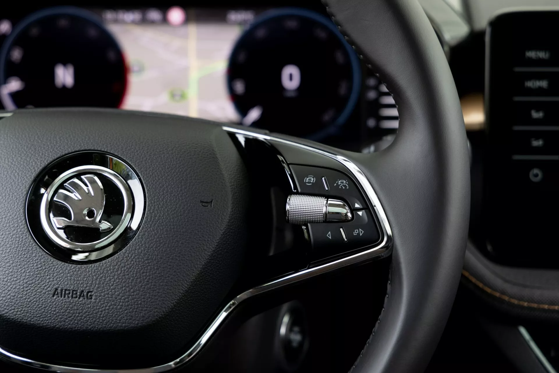 Detailní pohled na interiér z roku 2021 Škoda Fabia z pohledu řidiče, zaměřený na ovládací prvky na pravé straně volant. Kožený volant má stříbrný emblém s logem Škoda a pod ním vyražený nápis 'AIRBAG'. Ovládací panel na pravé straně obsahuje tlačítka pro multimediální systém, hlasitý telefon nebo hlasové příkazy, za volantem viditelné pádla. Rozostřené pozadí ukazuje panel přístrojů s otáčkoměrem a tachometrem a digitální displej s informací. Částečně viditelná je také středová konzole nebo multimediální systém na pravé straně.
