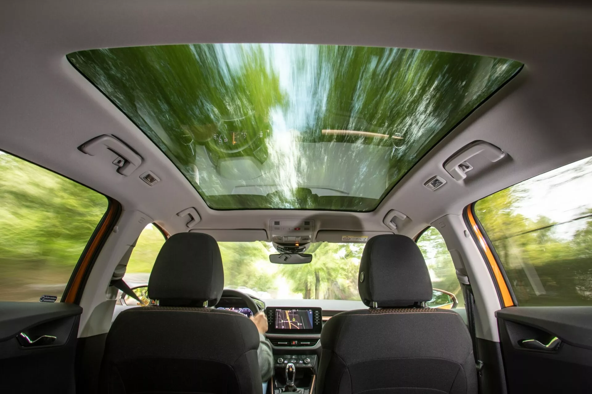 Pohled na interiér vozu Škoda Fabia 2021 z pohledu zadního sedadla, který ukazuje přední sedadla s tmavým čalouněním, panoramatické střešní okno, podsvícenou dotykovou obrazovku infotainmentu na palubní desce, ovládací panely, reproduktory integrované do dveří a ovládání oken, zpětné zrcátko s možnými high-tech funkcemi a horní konzolu pro další ovládání, což vše naznačuje moderní, pohodlný a technicky vylepšený zážitek z jízdy.
