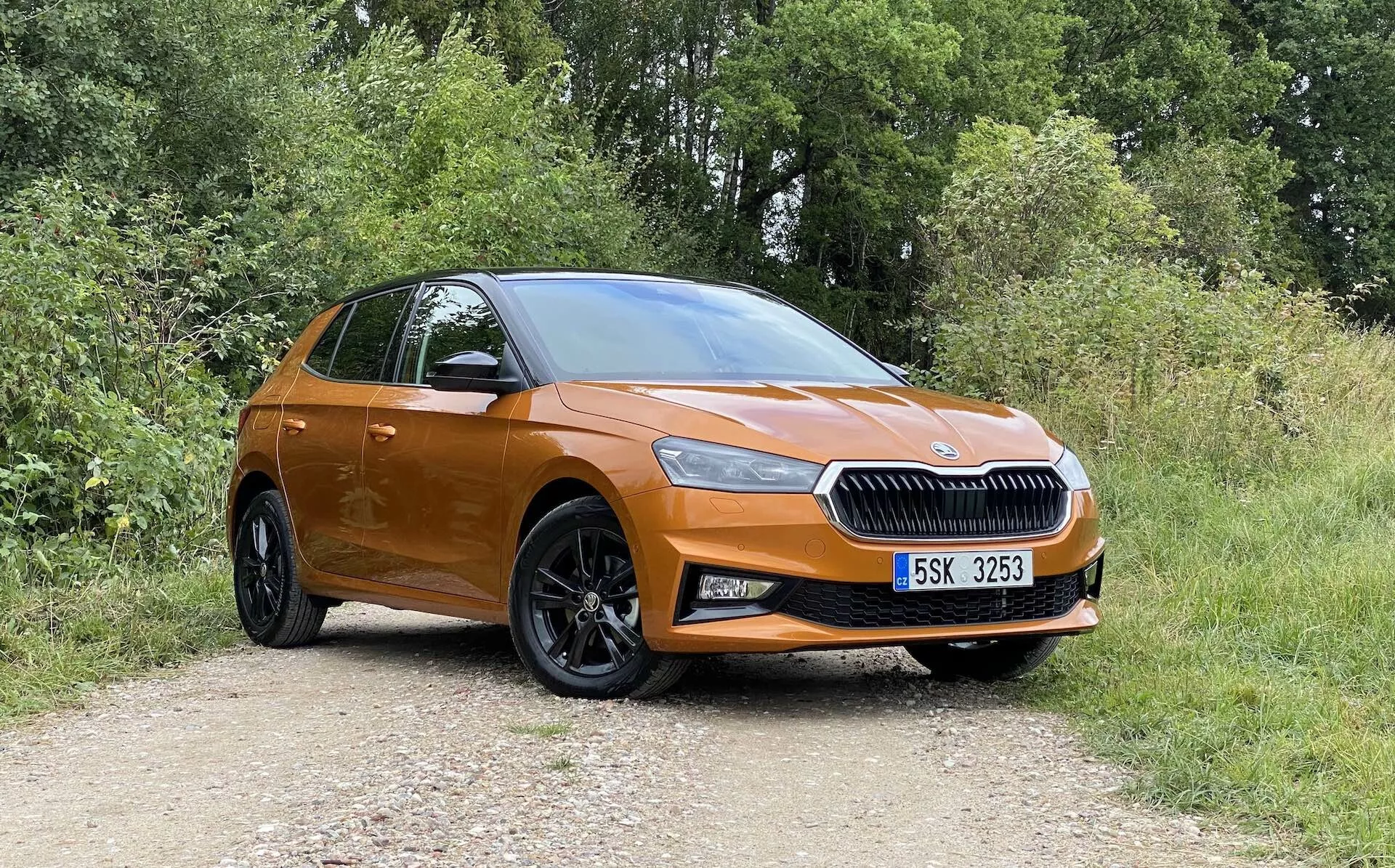 Plný boční profil moderního, oranžového hatchbacku Škoda Fabia 2021 zaparkovaného na nezpevněné cestě s vegetací v pozadí, zachycený mírně šikmo zepředu, zvýrazňující stranu řidiče. Mezi rysy patří výrazná mřížka s centrálním logem značky, elegantní světlomety, mlhová světla, velká černá vícepaprsková kola z lehké slitiny, barevně sladěné kliky dveří a zrcátka, klesající střecha a náznak zadního víka a světelné sestavy.