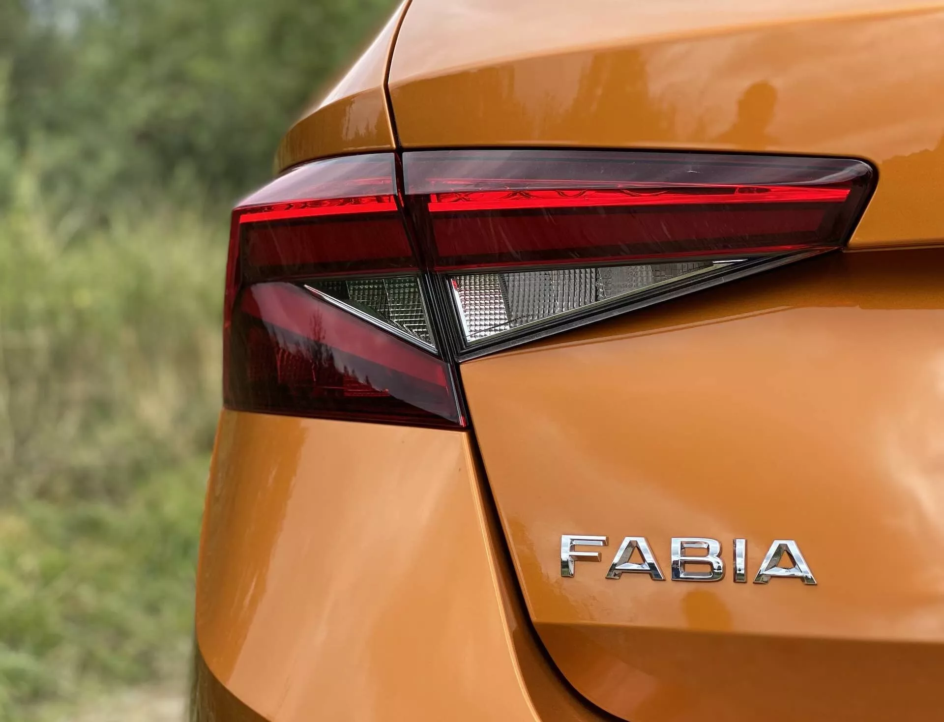 Zblízka záběr zadního rohu Škoda Fabia 2021 zaměřující se na ostrý, úhlový design zadního LED světla a výrazné logo 'FABIA' na lesklé, kovově oranžové karoserii. Hladké křivky a jemné vyklenutí kolem zadního světla dodávají moderní design, zachyceno z mírně vyvýšeného úhlu s malou hloubkou ostrosti.