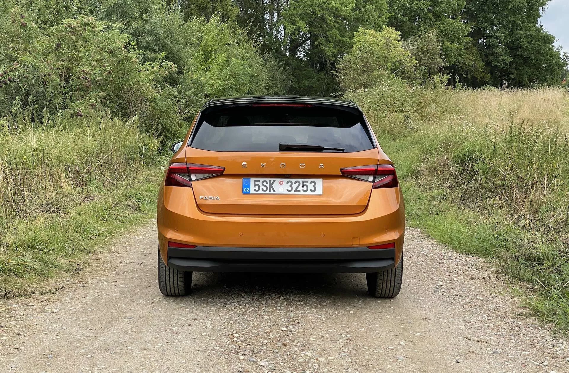 Zadní pohled na světle metalickou oranžovou Škodu Fabia 2021 hatchback fotografovanou za denního světla na úzké hliněné cestě v venkovském prostředí. Obrázek zachycuje moderní zadní design s nepřerušovanou zadní světelnou lištou, ostrými hranatými zadními světly, integrovanými odrazkami, stěračem zadního okna a zadním spoilerem pro aerodynamiku, zdůrazňující lesklý povrch auta a praktické funkce.