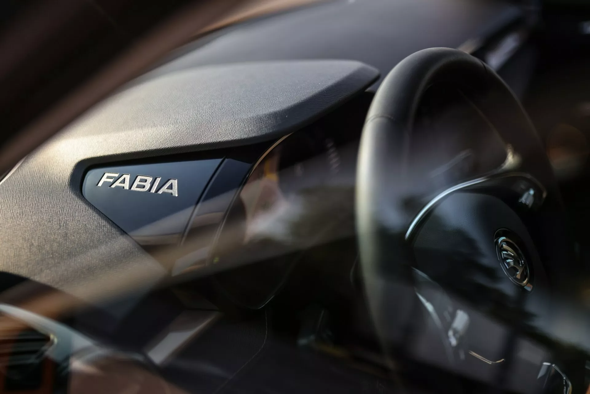 Detailní záběr interiéru vozu Škoda Fabia 2021, zaměřený na pravou stranu přístrojové desky. Výrazně je zobrazen nápis 'FABIA', zatímco na levé straně je částečně vidět moderní volant s logem Škoda. Přístrojová deska má zrnitou texturu a mělká hloubka ostrosti zdůrazňuje značku s jemně neostrým pozadím.