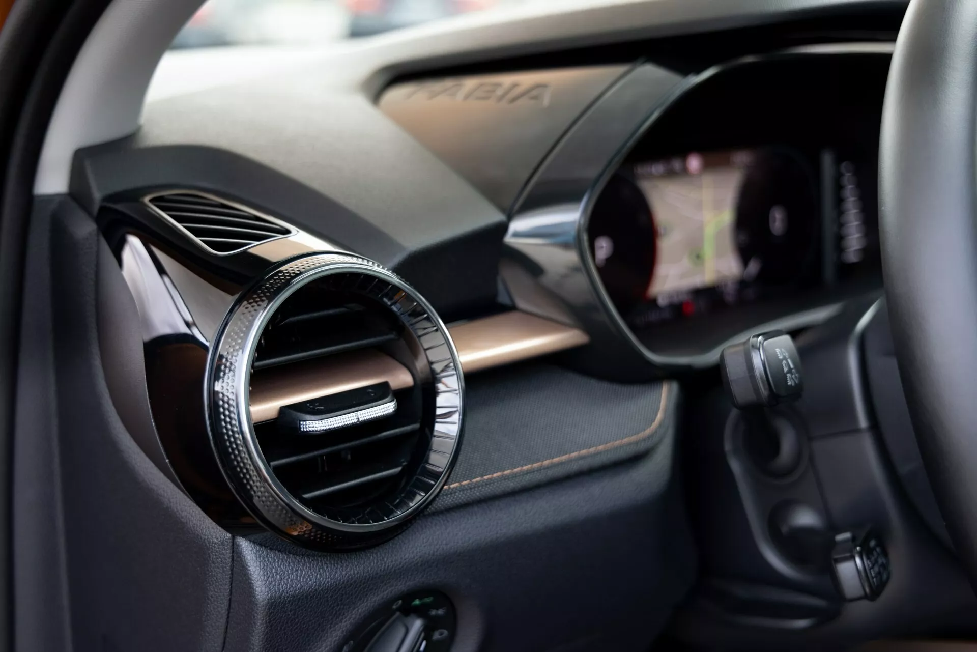 Detailní pohled na interiér Škoda Fabia 2021 zaměřený na stranu spolujezdce. Obrázek zachycuje kulatý ventilační otvor s kovovým nebo chromovým povrchem proti panelu s texturou připomínající uhlíková vlákna, což naznačuje sportovní nebo vyšší výbavu. Vpravo je viditelný okraj dotykové obrazovky infotainmentu integrované do palubní desky. Pod ventilačním otvorem jsou různá ovládací tlačítka, nejspíš pro audiosystém, klimatizaci nebo jiné funkce vozidla. Malá hloubka ostrosti upozorňuje na ventilační otvor a jemně naznačuje okolní technologické komponenty.