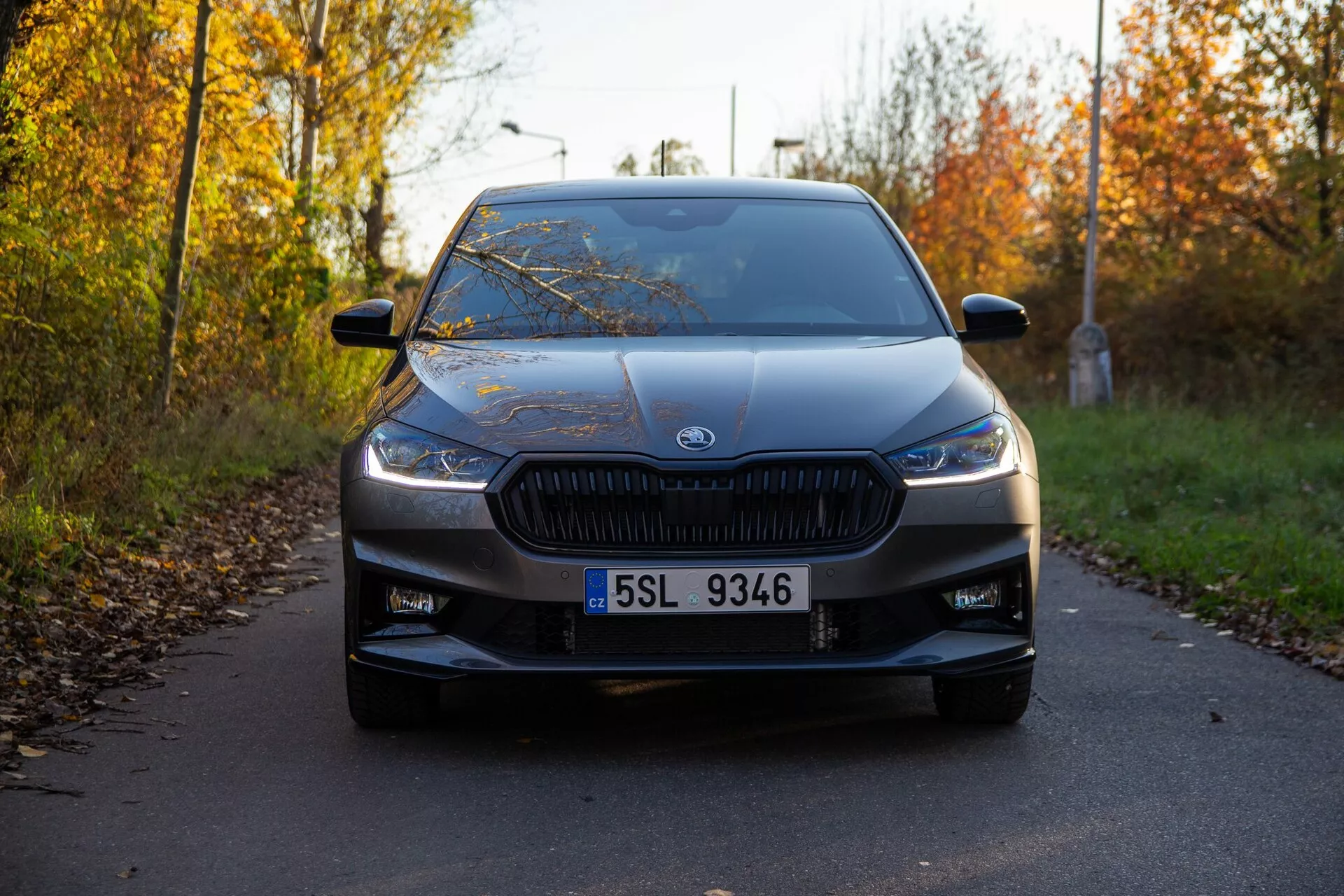 Pohled zepředu na Škoda Fabia 2021 zachycený z nízkého úhlu, zdůrazňující charakteristickou mřížku s vertikálními žebry a moderními, úzkými světlomety. Auto, umístěné ve středu rámu, je zaparkované na úzké cestě lemované podzimním listím, ozdobené výrazným logem výrobce umístěným ve středu mřížky. Viditelné jsou další designové prvky jako čitelná SPZ, nasávací otvory na spodním nárazníku, jemné obrysové linie na kapotě, rozsáhlé čelní sklo a boční zrcátka s ukazateli. Měkké osvětlení a dlouhé stíny naznačují, že obraz byl pořízen buď časně ráno nebo pozdě odpoledne a auto je ve vynikajícím stavu.