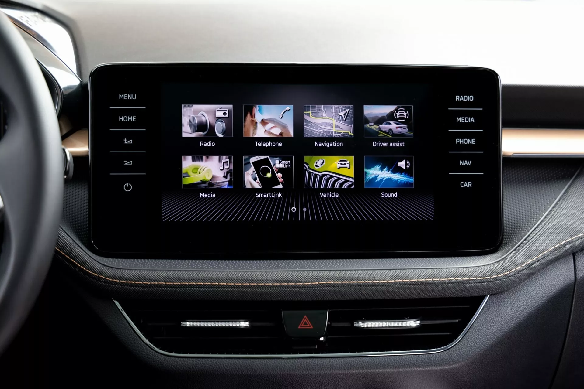 Detailní pohled na dotykový infotainment systém modelu Škoda Fabia 2021 z perspektivy cestujícího nebo řidiče v přední řadě, zobrazující elegantní středovou konzolu rámovanou kartáčovaným hliníkem. Displej ukazuje uživatelsky přívětivé grafické rozhraní s barevnými ikonami pro Domů, Rádio, Telefon, Navigaci a další. Pod obrazovkou je řada fyzických tlačítek a možný CD slot přidávající multifunkčnost. Prémiové čalounění s kontrastním prošitím naznačuje luxusnější interiérový design.