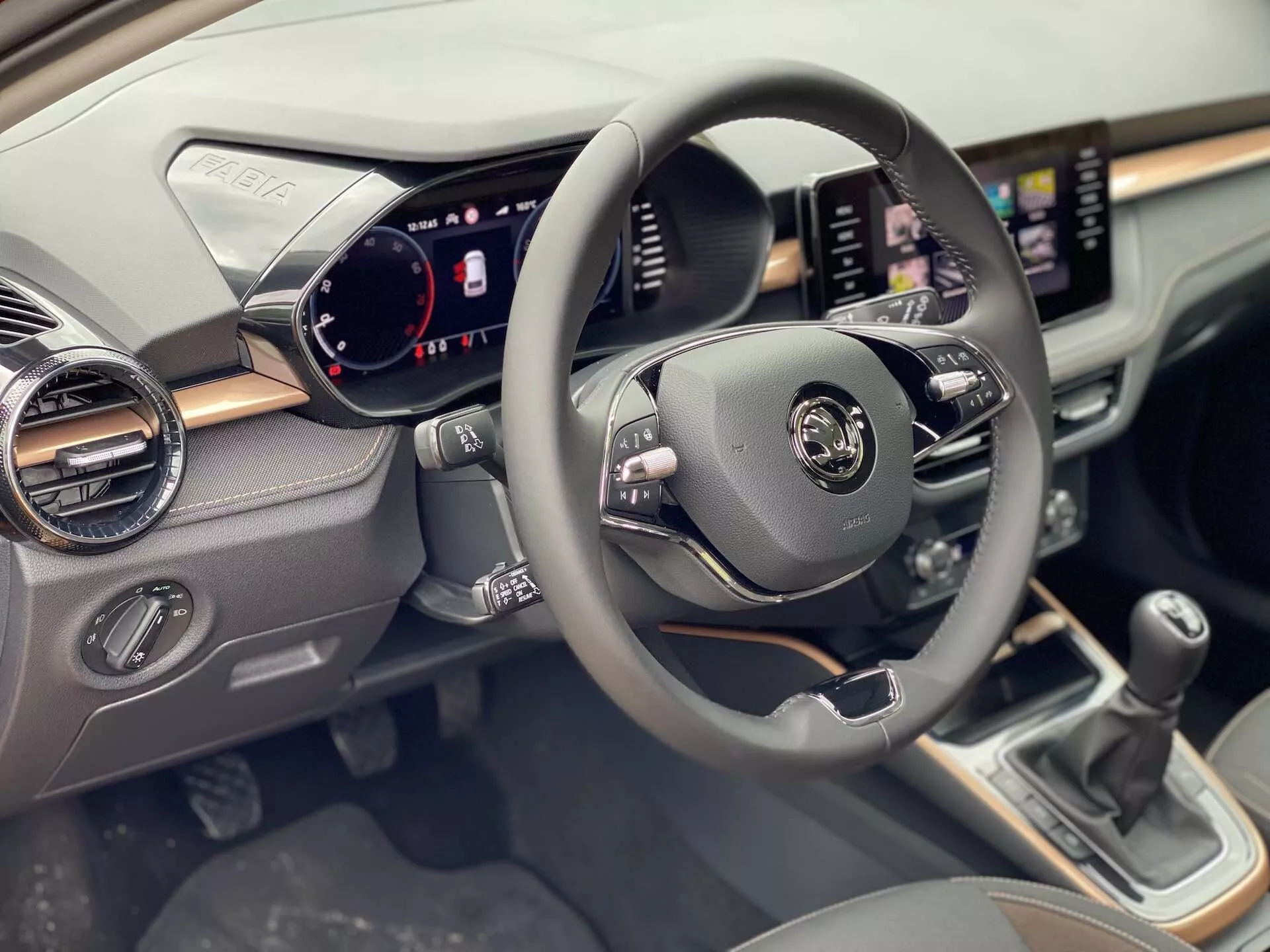 Pohled do interiéru Škody Fabia 2021 zaměřený na stranu řidiče, zobrazující středový volant s multifunkčními ovládacími prvky pro audio, hlasové ovládání a tempomat. Digitální přístrojová deska zobrazuje tachometr, rychloměr a indikátory stavu vozidla. Na palubní desce se nachází dotykový infotainment systém s funkcionalitami pro navigaci a média. Pod obrazovkou jsou ovládací prvky pro klimatizaci. Na středové konzoli je vidět řadicí páka manuální převodovky.