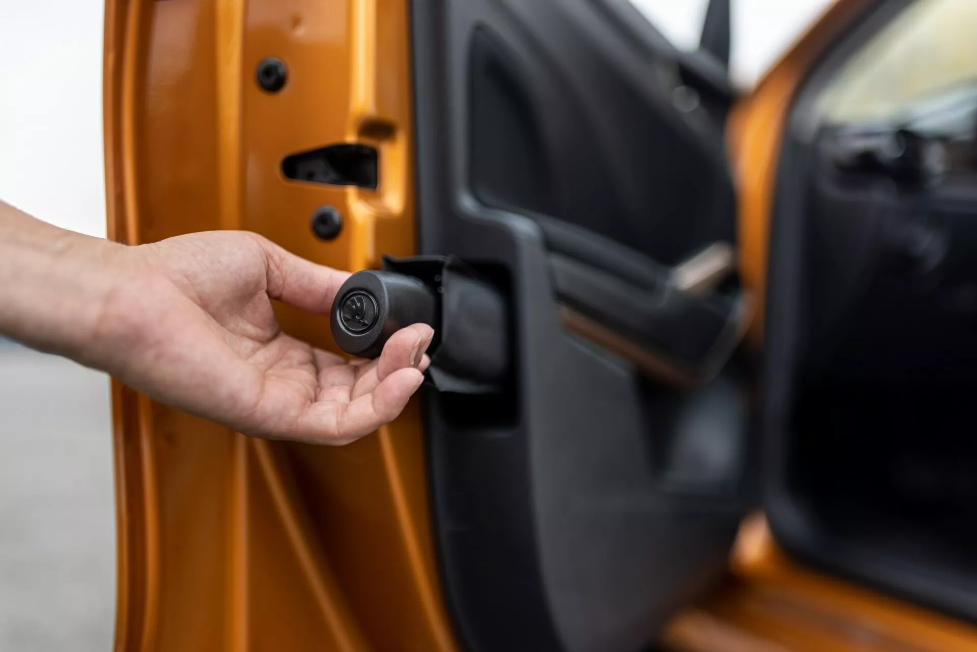 Detailní záběr na dveře řidiče vozu Škoda Fabia 2021, mírně pootevřené, pohled z exteriéru s viditelným vnitřním okrajem. Ruka drží kompaktní černý, kruhový klíč, který označuje bezklíčový vstup nebo systém zabezpečení auta. Auto je jasně oranžové s metalickým povrchem, což ukazuje na pokročilé zamykací mechanismy a personalizované bezpečnostní prvky.