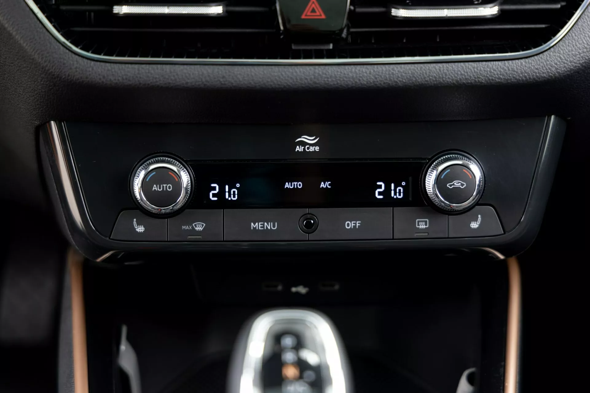 Zblízka pohled na ovládací panel klimatizace vozu Škoda Fabia 2021 umístěný na středové konzoli, s duálními teplotními ovladači s digitálními displeji se zobrazením '20', integrovanými tlačítky pro automatické ovládání klimatizace ('AUTO'), klimatizaci ('AC'), nastavení menu ('MENU') a vypnutí systému ('OFF'). Okolní lesklý černý rámeček obklopuje informační displej nad ovladači s viditelným tlačítkem výstražného trojúhelníku (ikona trojúhelníku) nahoře. Panel je umístěn v prémiovém matném a dřevěném interiéru, který zdůrazňuje moderní design a technologii.