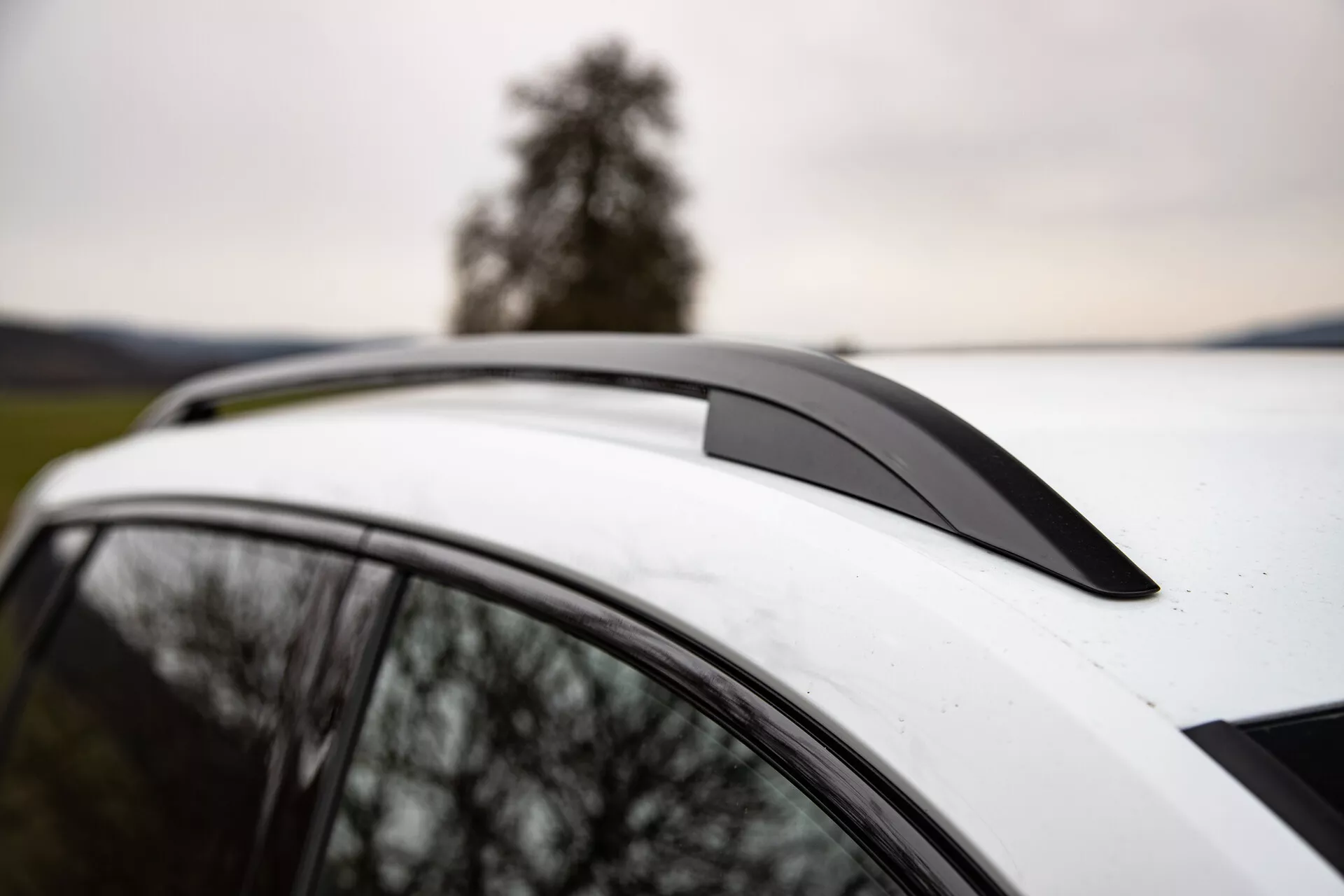 Detailní pohled na Škoda Karoq 2021 zobrazující spoiler na střeše, bílou střechu s panoramatickým střešním oknem a integrované střešní nosiče na pozadí stromu a zamračené oblohy, zdůrazňující aerodynamický design vozidla, luxusní prvky a možnosti využití.