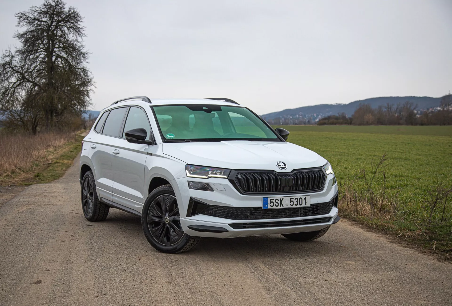 Bílé SUV Škoda Karoq 2021 zaparkované na venkovské prašné cestě, předvádějící pohled na přední levou čtvrt včetně mřížky, světlometů a designu kol, s důrazem na moderní prvky jako LED osvětlení a parkovací senzory.