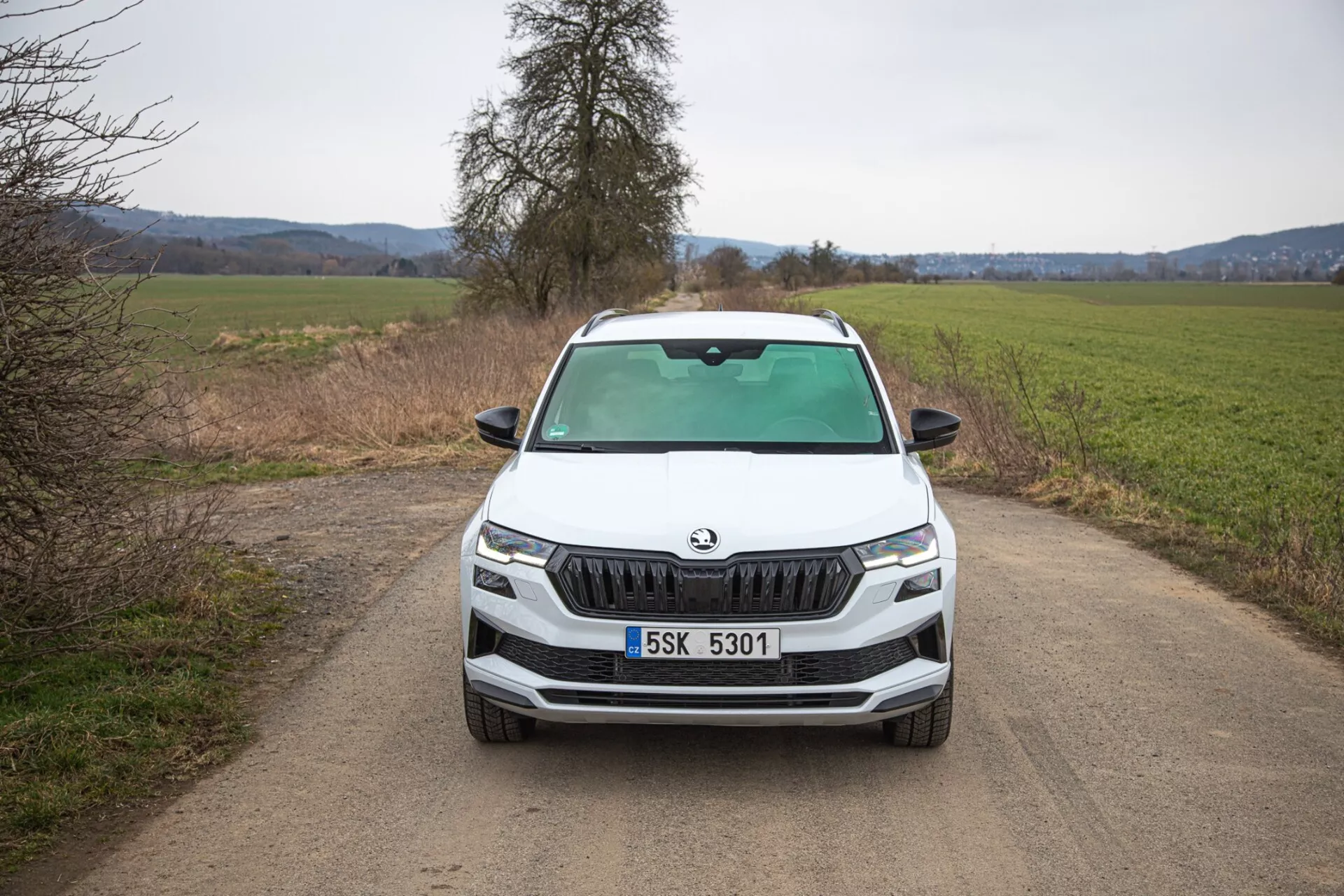 SUV Škoda Karoq 2021 zobrazující přímý čelní pohled na nezpevněné úzké venkovské cestě, zdůrazňující charakteristickou Škoda mřížku, ostrá LED světlomety, mlhová světla v černém lemování pro možné terénní schopnosti, dynamické linie kapoty a pokročilé technologické prvky na přední části vozu.