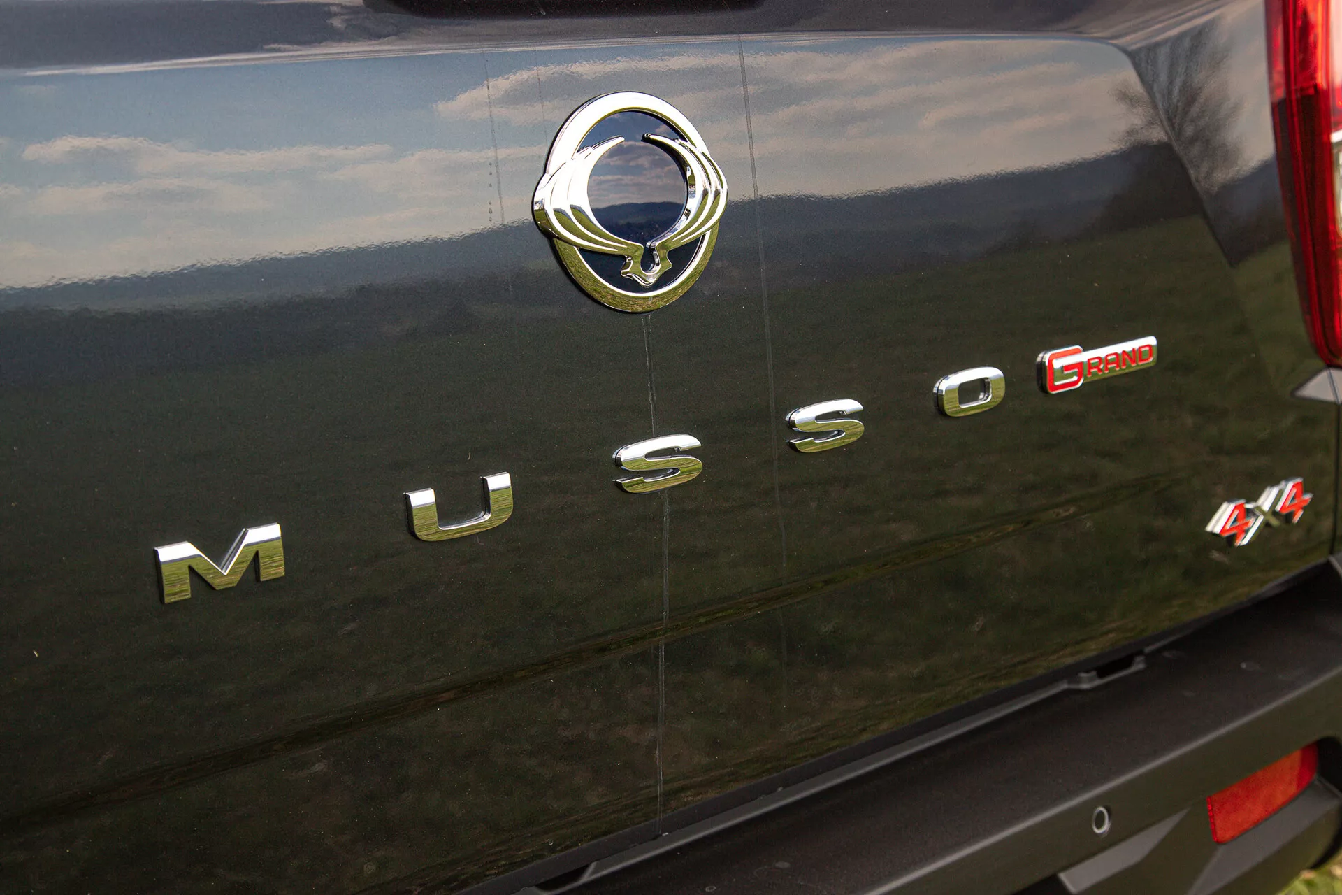 Detailní pohled na zadní část SsangYong Musso Grand z roku 2022 s chromovým nápisem ‘MUSSO’, stříbrnou emblémou výrobce se stylizovaným motivem ptáka a červenou značkou ‘E-XDI Sport’ udávající typ motoru a úroveň výkonu, umístěnou na tmavém, možná černém, zadním exteriéru vozidla s reflexními chromovanými a texturovanými detaily.
