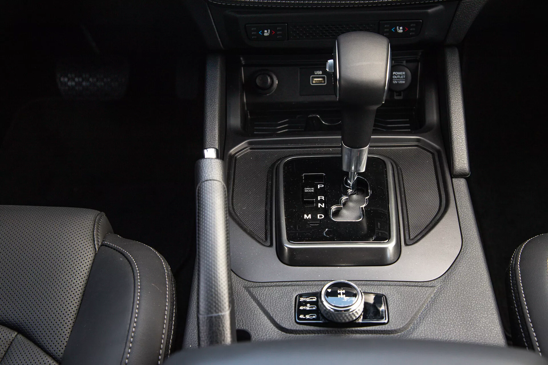 Detailní pohled na středovou konzolu SsangYong Musso 2022 s automatickou převodovkou, elektronickou parkovací brzdou, tlačítkem start/stop, přepínačem systému automatického start-stop, držáky na nápoje, aux vstupy, a viditelnými sedadly potaženými kůží, které zdůrazňují moderní design a luxusní prvky