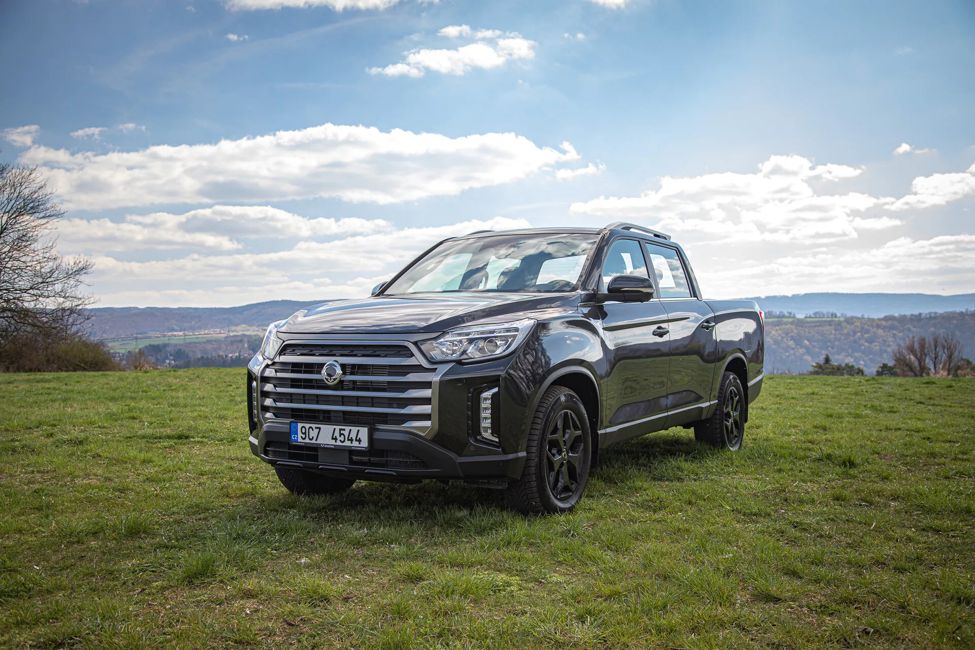 Pick-up SsangYong Musso Grand z roku 2022 zaparkovaný na travnaté krajině pod zamračenou oblohou, předvádějící svůj robustní design a schopnost jízdy v terénu z pohledu ze tří čtvrtin zepředu během dne.
