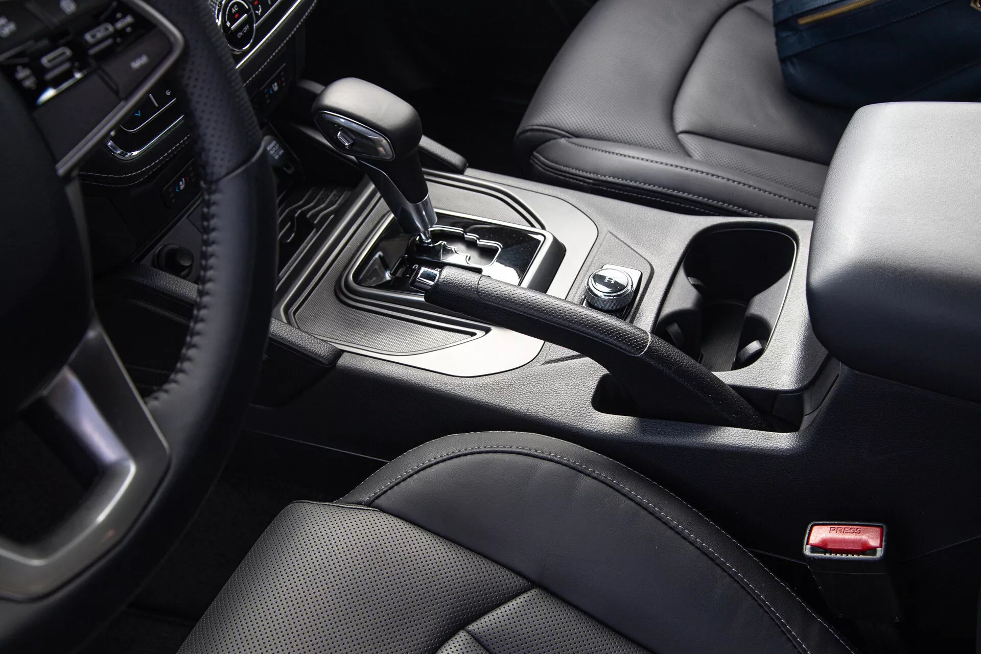 Pick-up SsangYong Musso Grand 2022 s pohledem shora a mírně zkoseným úhlem na středovou konzoli s voličem automatické převodovky v pozici 'Park', obklopeným ovladači infotainmentu a jízdních režimů, loketní opěrkou potaženou kůží a akcenty s designovým kovovým povrchem.