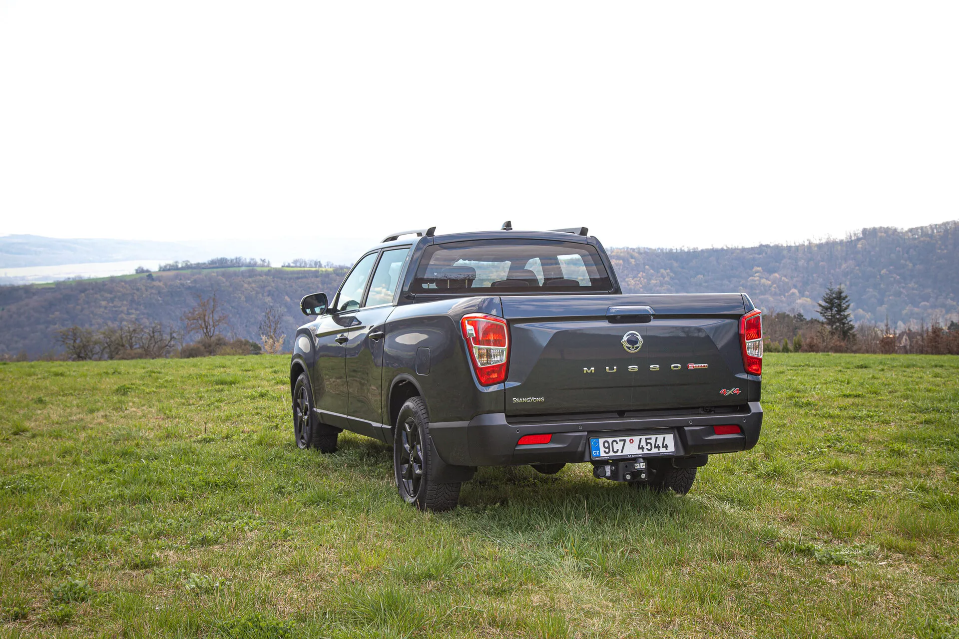 SsangYong Musso Grand pickup z roku 2022 zachycený z zadního úhlu, který zdůrazňuje jeho sportovně užitkový vzhled, čtyřdveřový design a venkovní všestrannost proti malebnému kopcovitému a říčnímu pozadí během dne