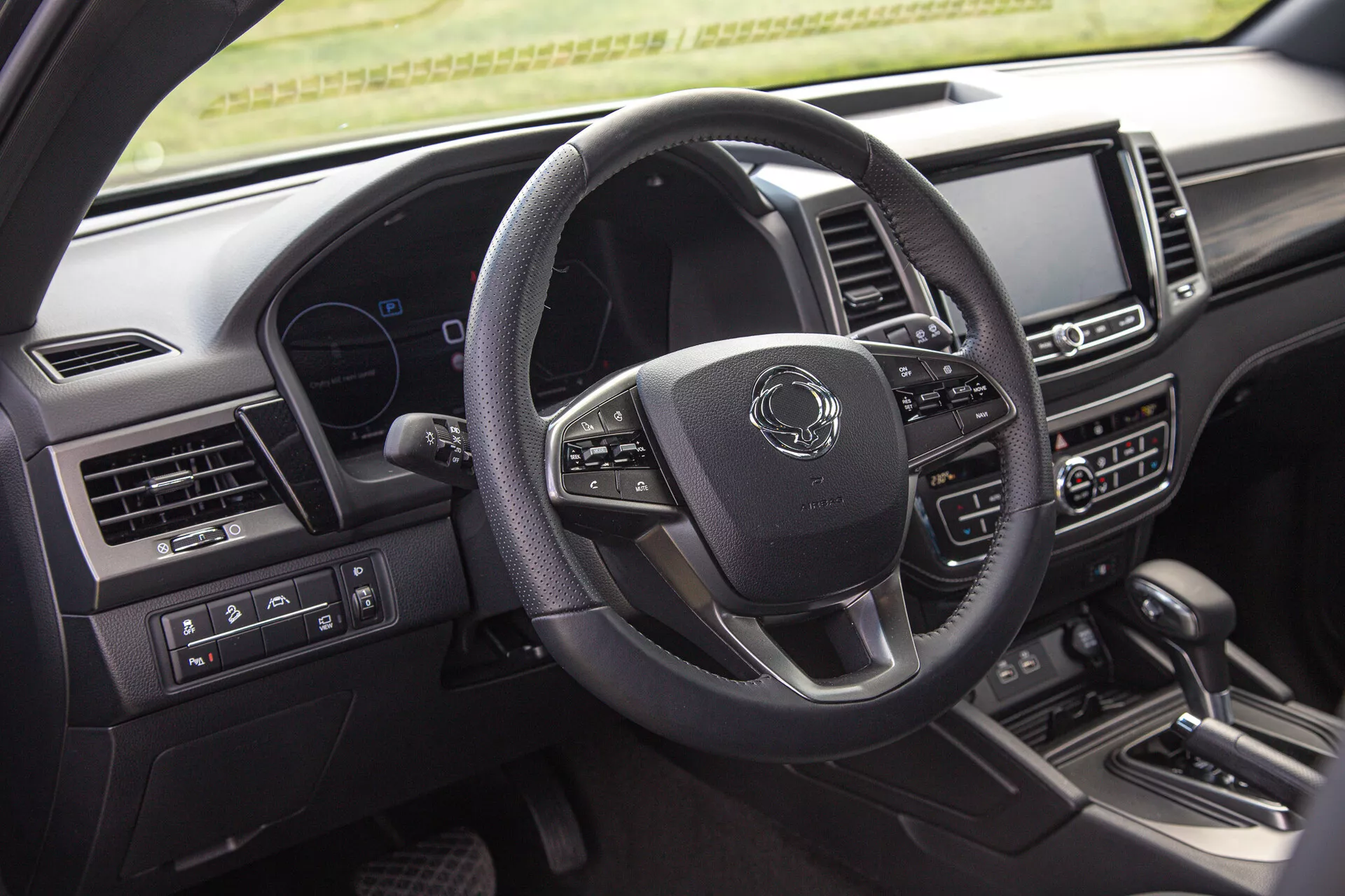 Interiérový snímek SsangYong Musso 2022 ukazuje stranu řidiče včetně moderního volantu se třemi rameny s multifunkčními tlačítky, digitálního přístrojového panelu a centrálního infotainment systému na palubní desce z pohledu zadního pasažéra
